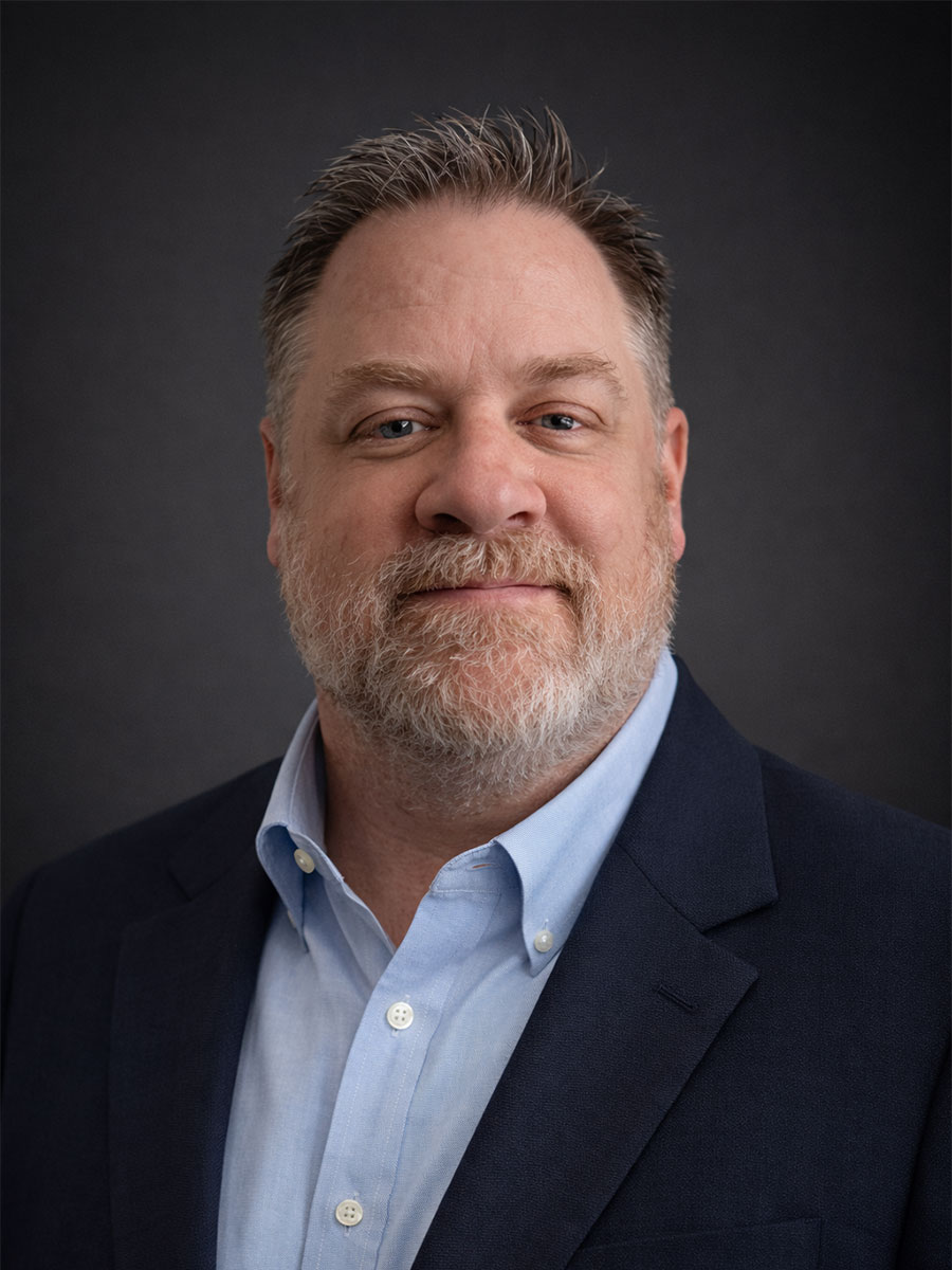 Middle-aged man with short gray hair and beard wearing a navy blazer and light blue shirt against a dark background.