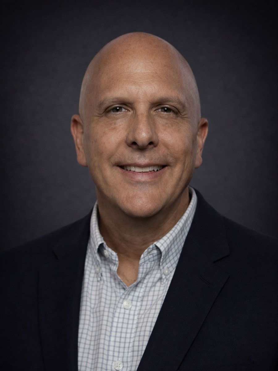 Bald man wearing a dark blazer and checked shirt smiles in front of a dark, plain background.