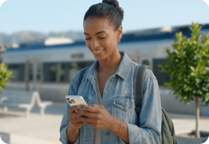 Smiling woman in denim jacket using smartphone outdoors with a blurred train and trees in the background.