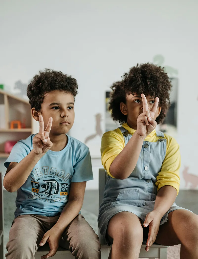 Two children sitting side by side, each holding up two fingers in a peace sign.