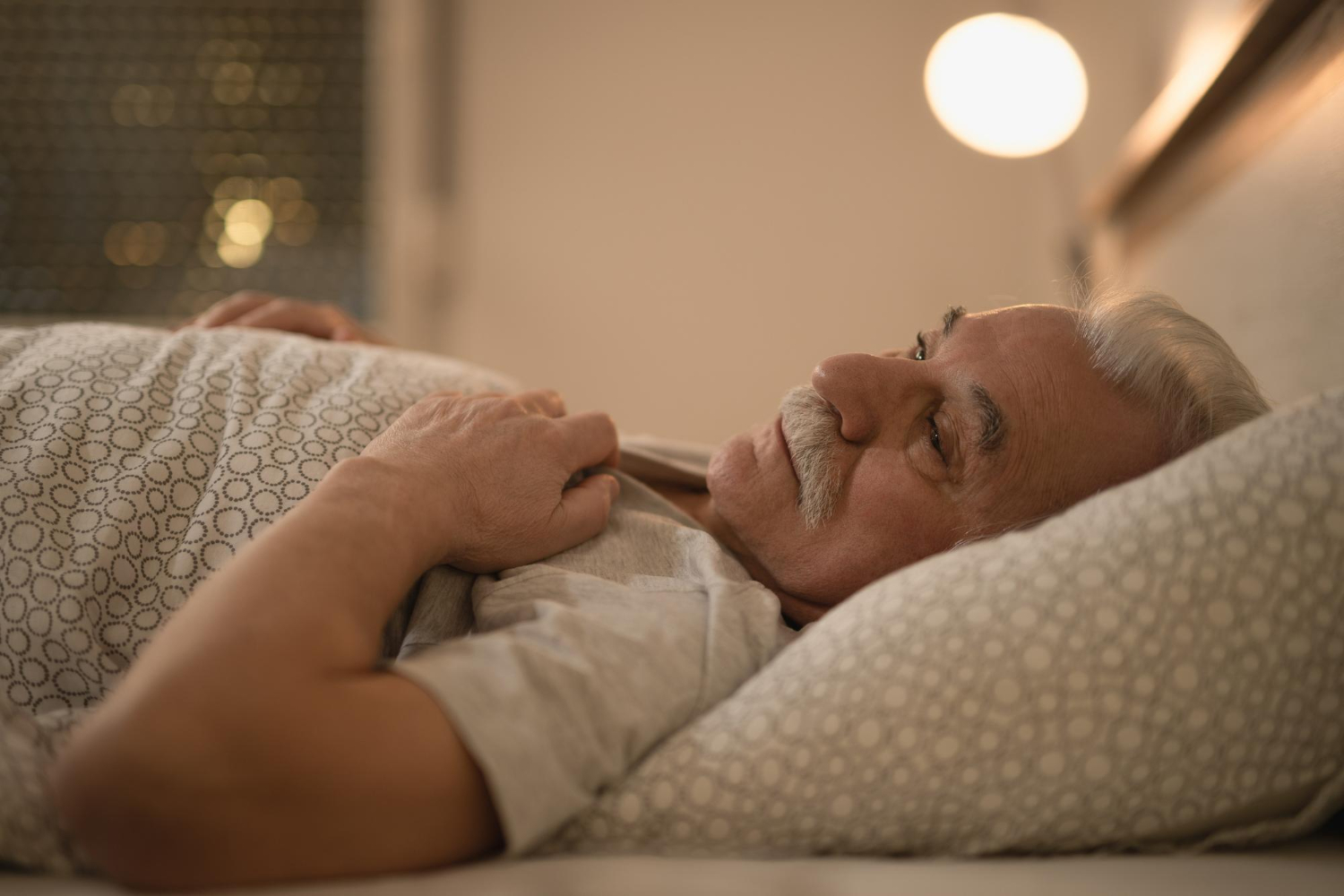 Elderly man lying in bed with hands resting on chest, looking contemplative in warm bedroom lighting.
