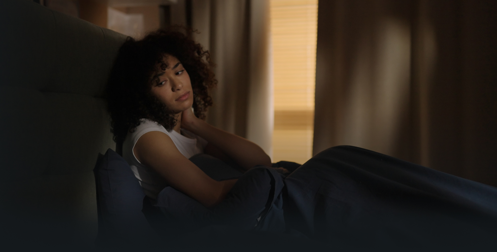 Woman sitting in bed with a dark blanket, resting her hand on her neck and looking concerned.