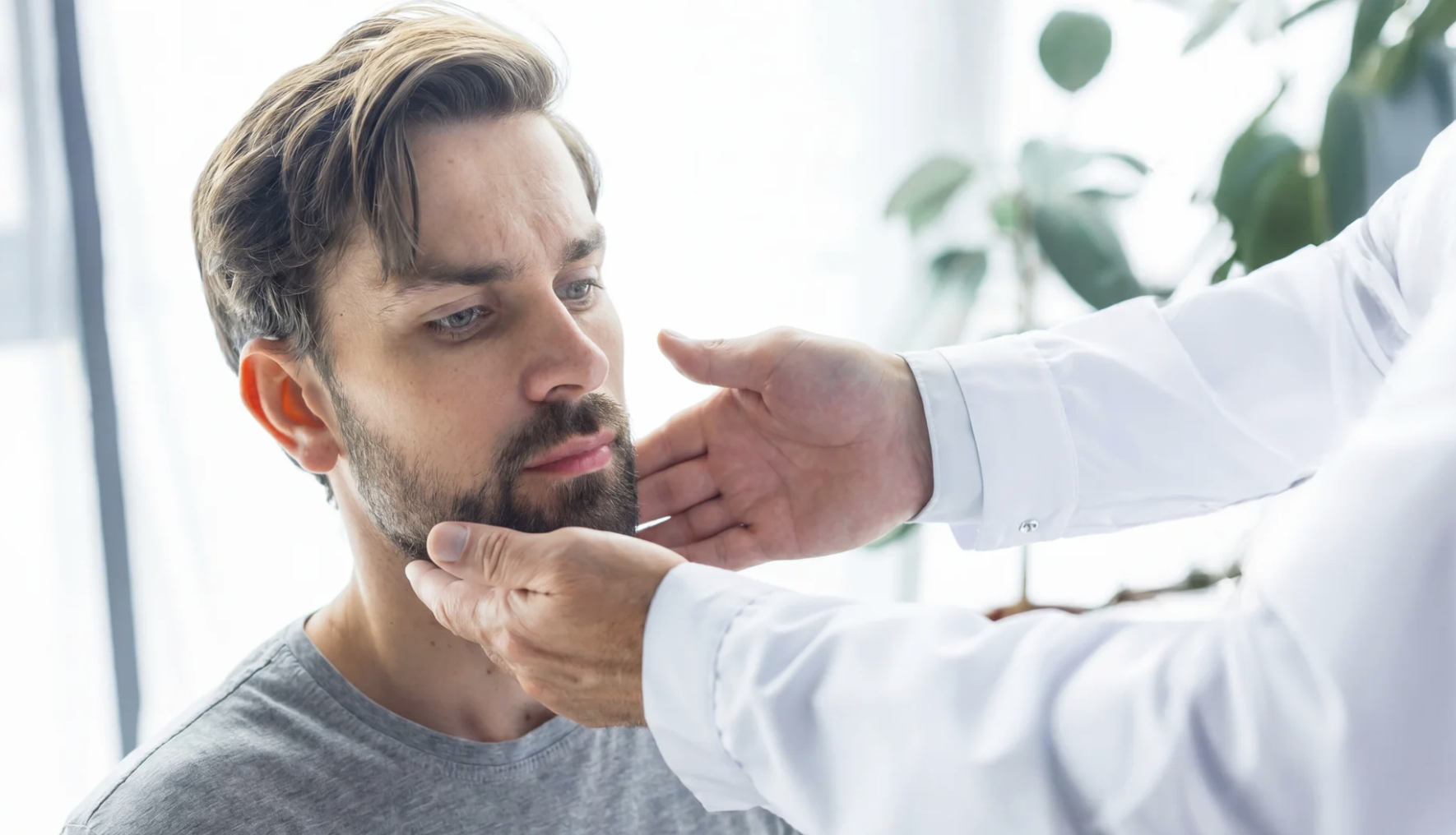 Dentist in white coat using both hands to palpate a patient's jaw during a clinical exam for TMJ disorder