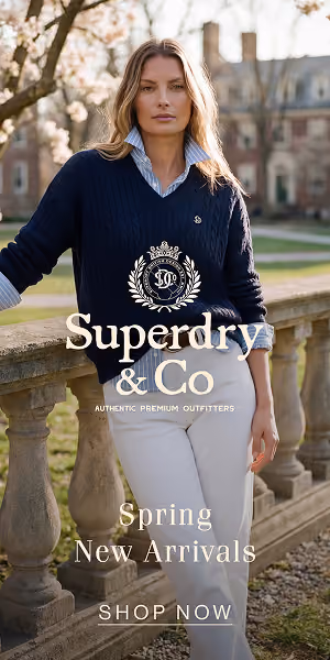 Woman wearing navy sweater and white pants standing by stone railing with building and trees behind.