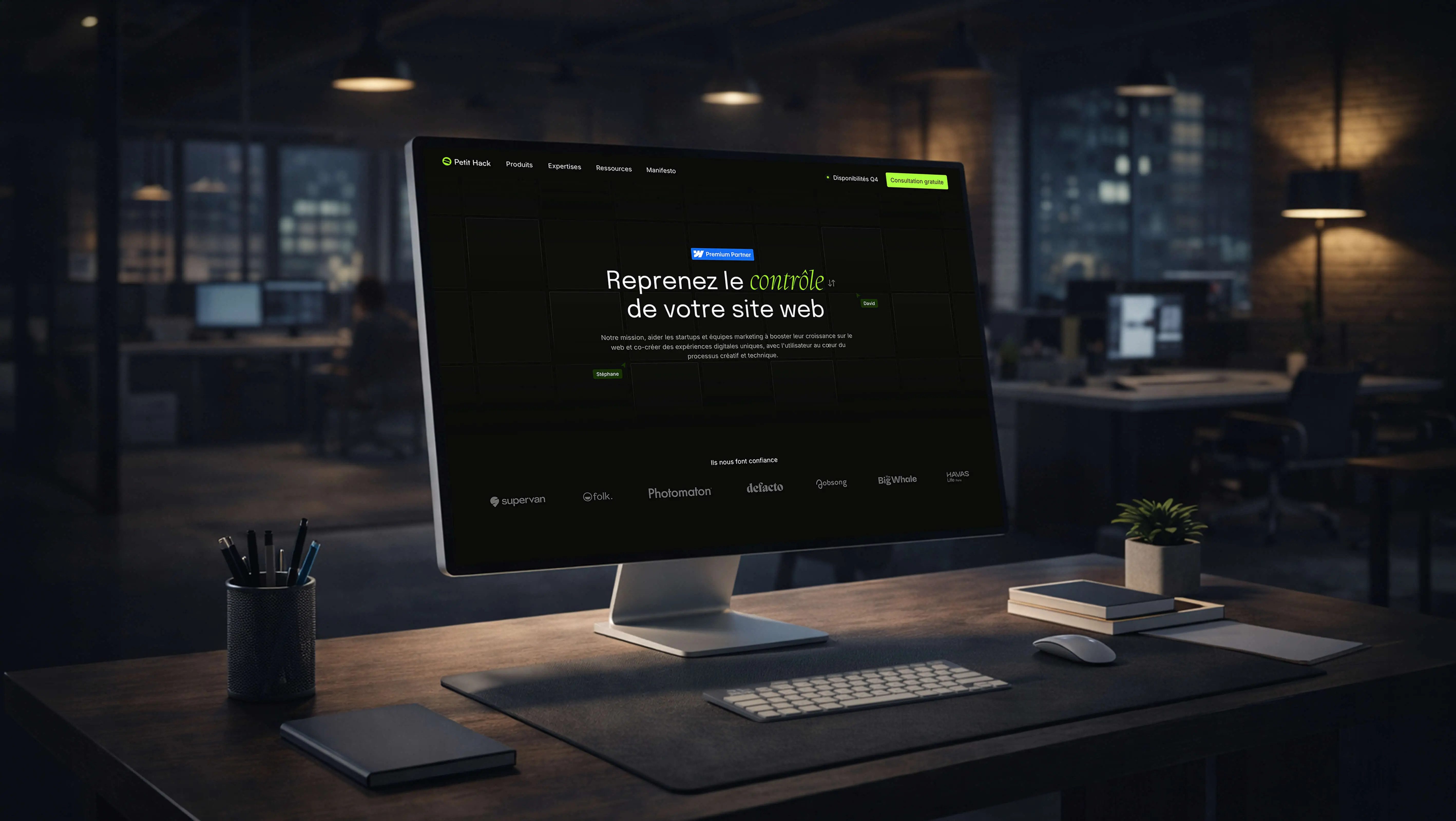 Mockup du site Petit Hack affiché sur un écran dans un bureau sombre, avec une interface dark mode et des logos de clients visibles à l'écran