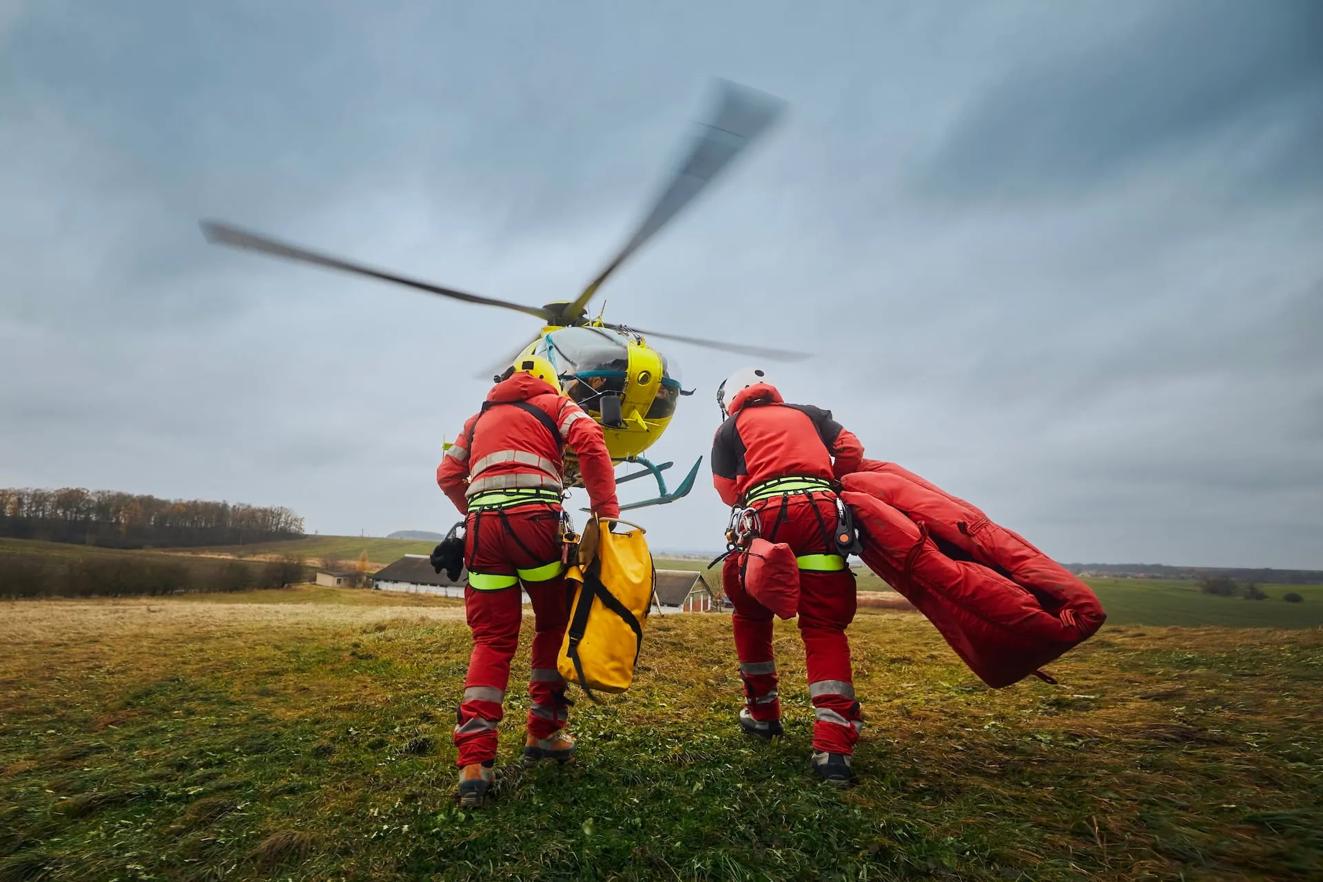Two paramedics in red suits carrying gear toward a yellow rescue helicopter