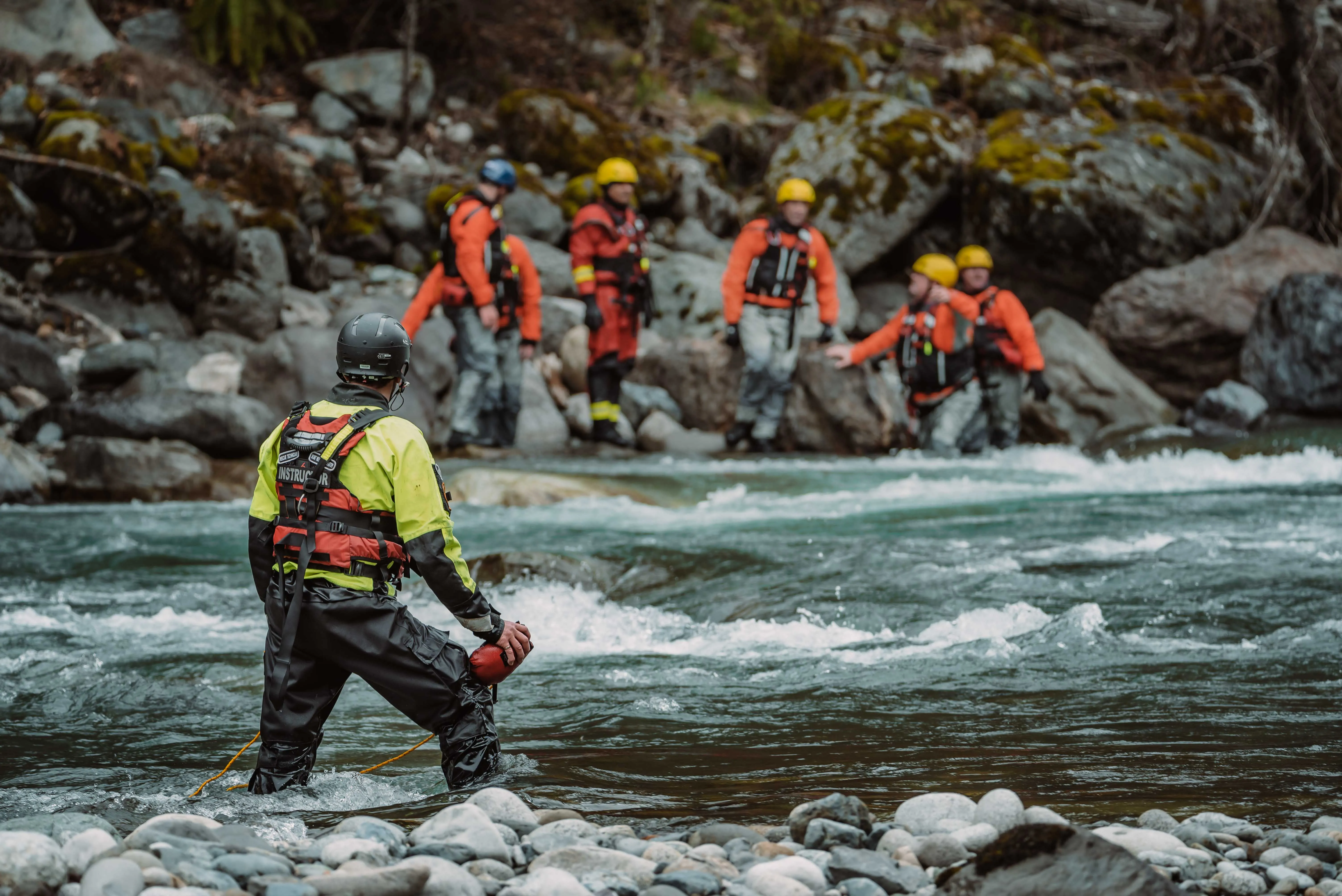 Swiftwater rescue instructor watching students in helmets and drysuits cross a river