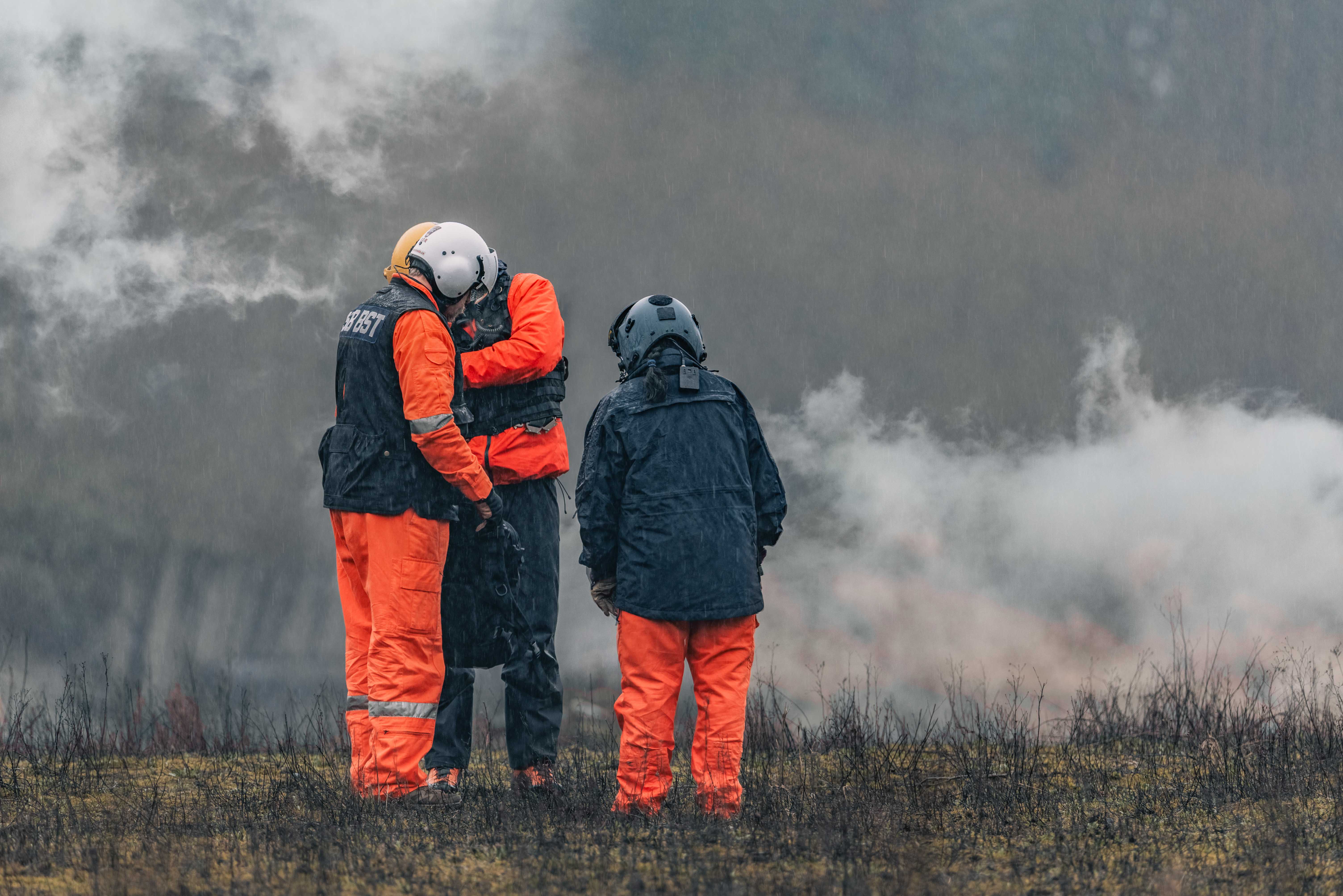 Three rescue team members in orange suits and helmets talking in a smoky field