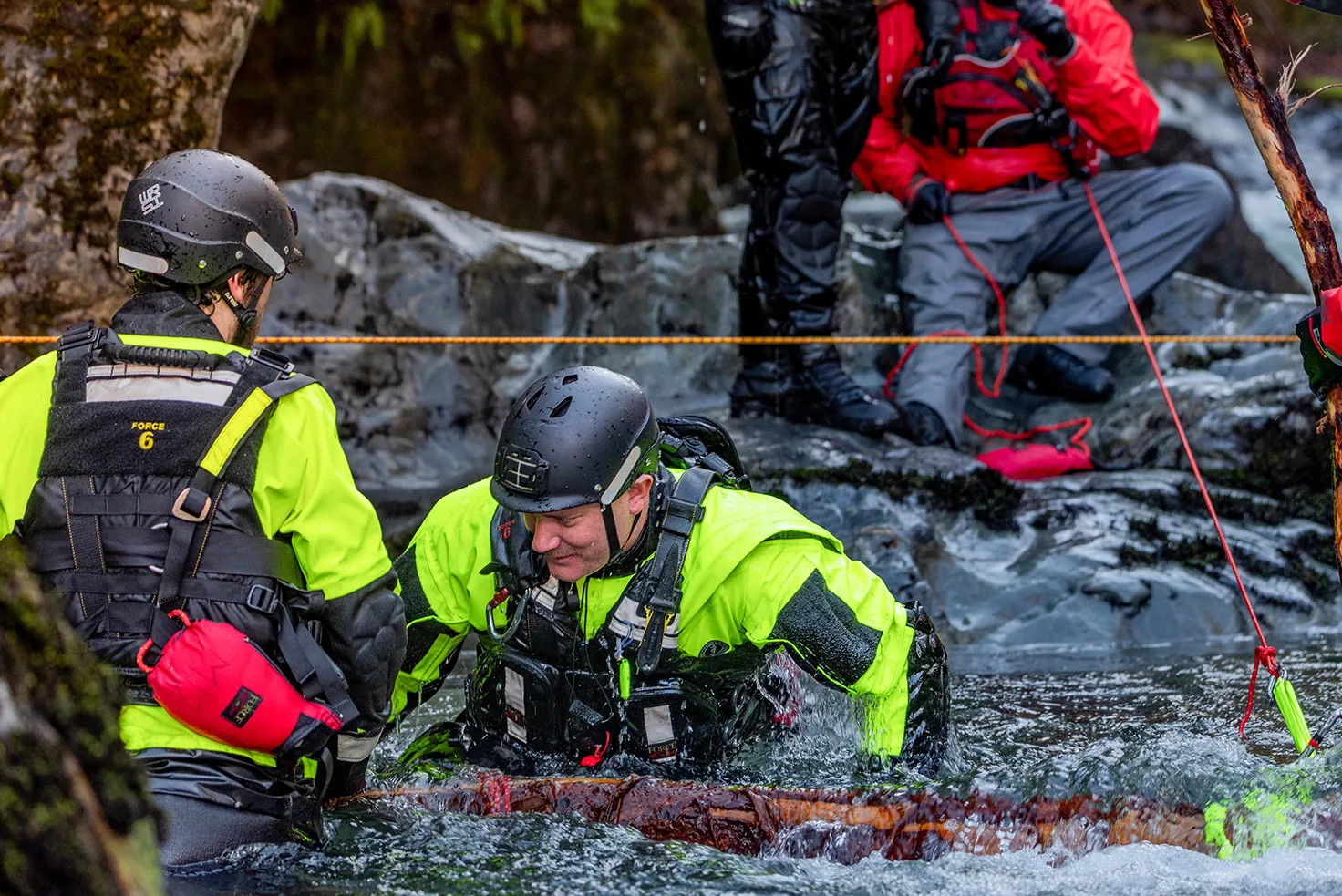 Swiftwater rescue technicians in helmets and drysuits working beside a log in a fast creek