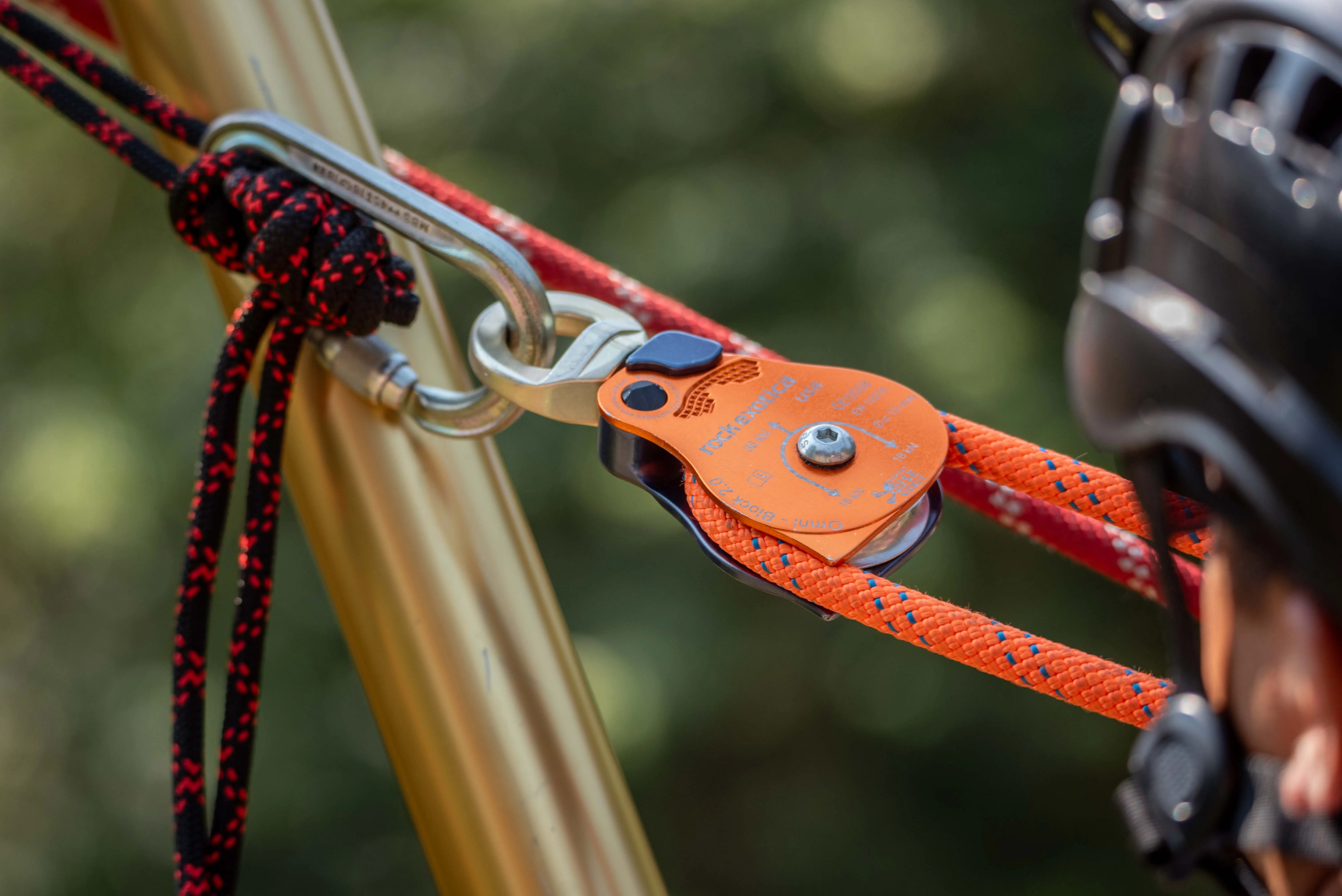 Orange rescue pulley and carabiners rigged to a tripod leg beside a helmeted rescuer