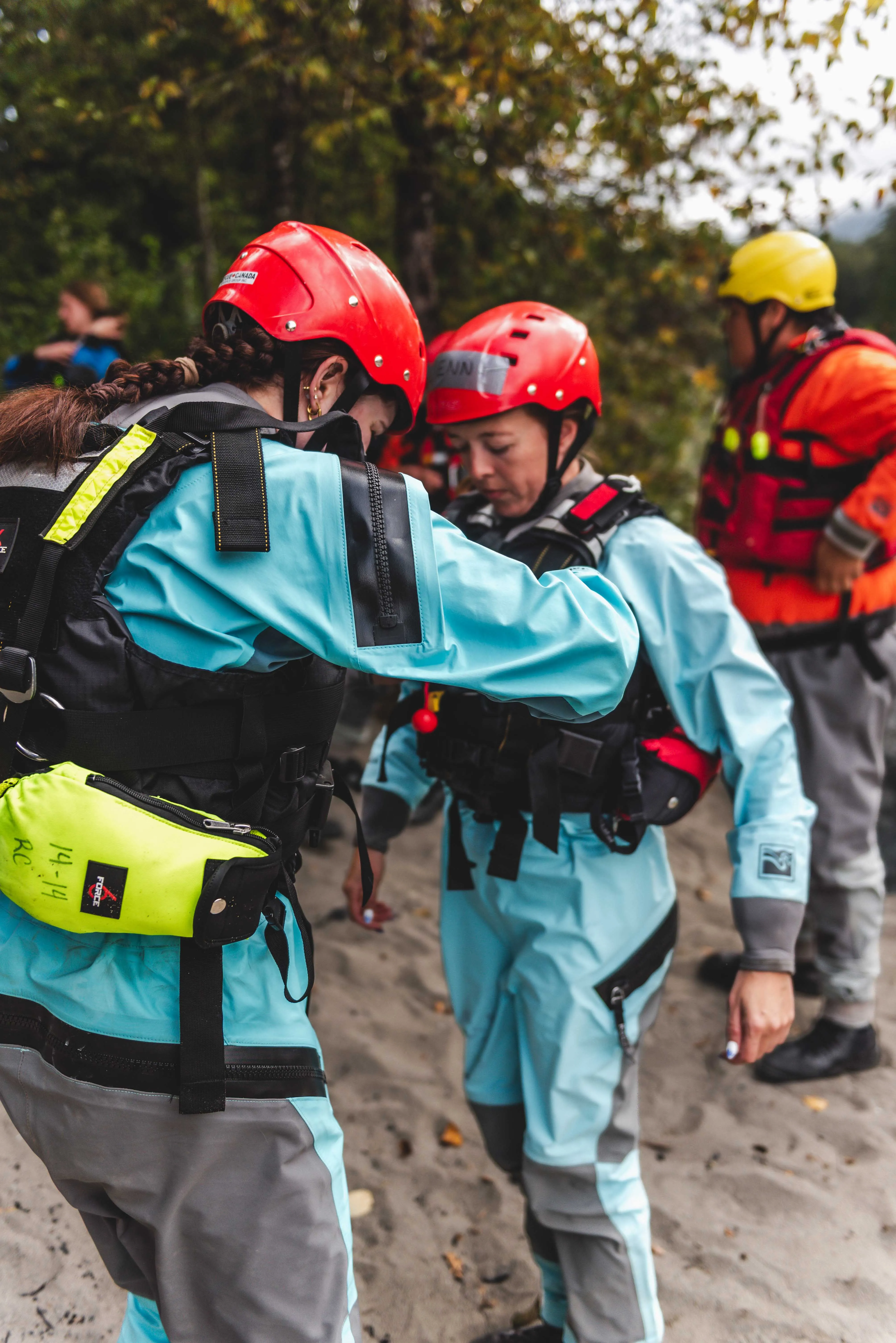 Swiftwater rescue students in red helmets and drysuits checking each other's gear on a riverbank