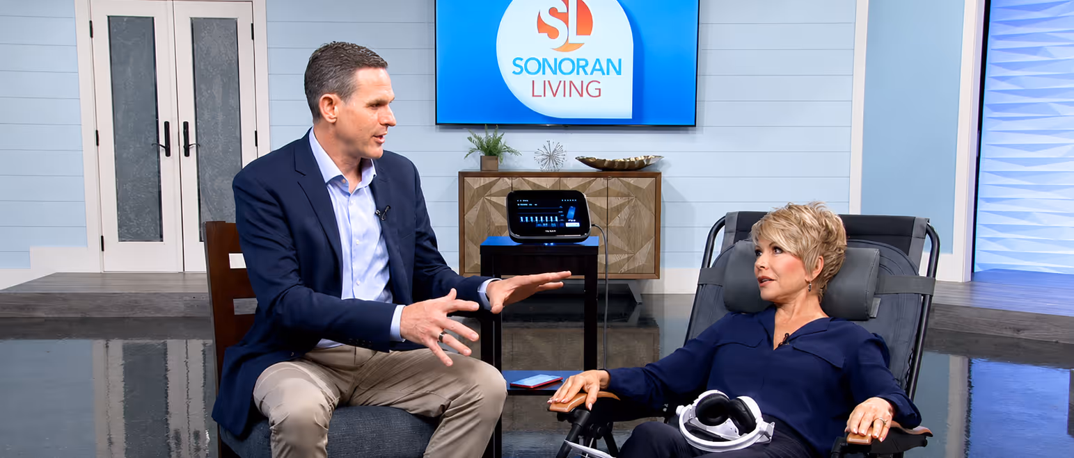 Man in blazer talks to woman seated in reclining chair with headset, Sonoran Living logo on screen.