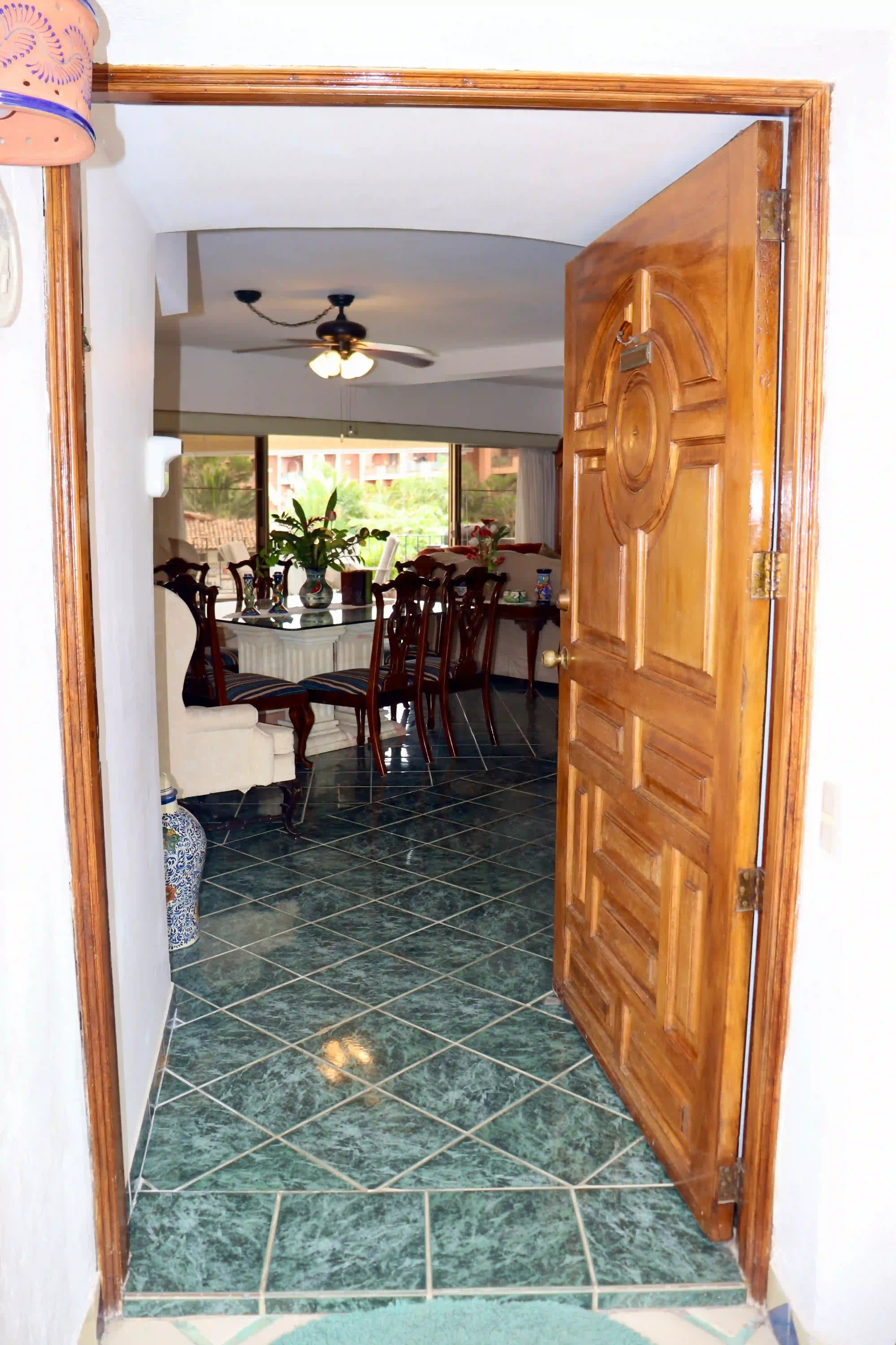 Bright entrance patio of the Royal Pacific 2304 condo in Marina Vallarta, featuring a solid wood front door, golden-framed windows, and a potted green plant against clean white walls.