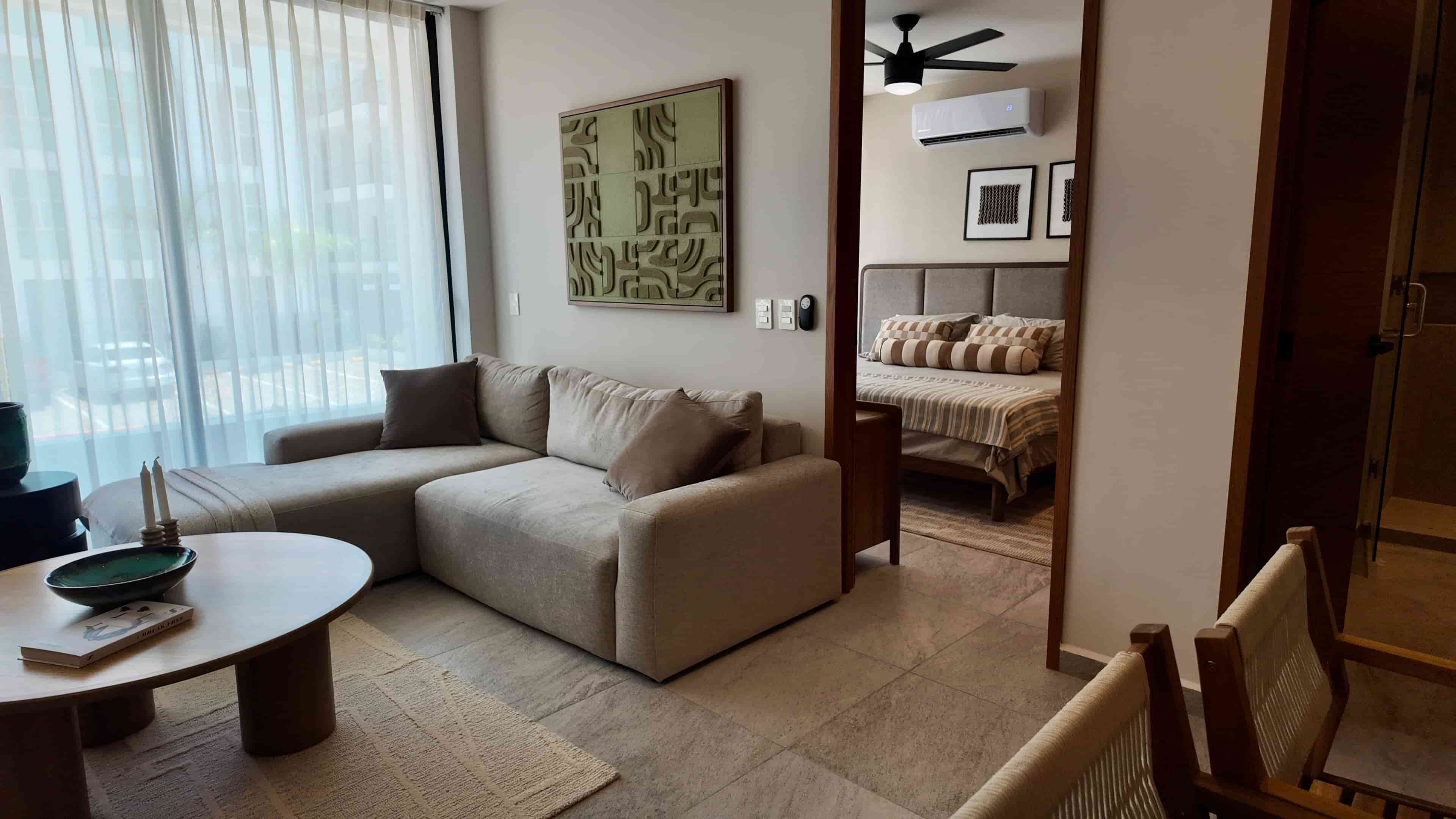 Comfortable living room layout with an L-shaped sofa and round coffee table, looking into the adjacent bedroom area of the TERRA 242 model unit.
