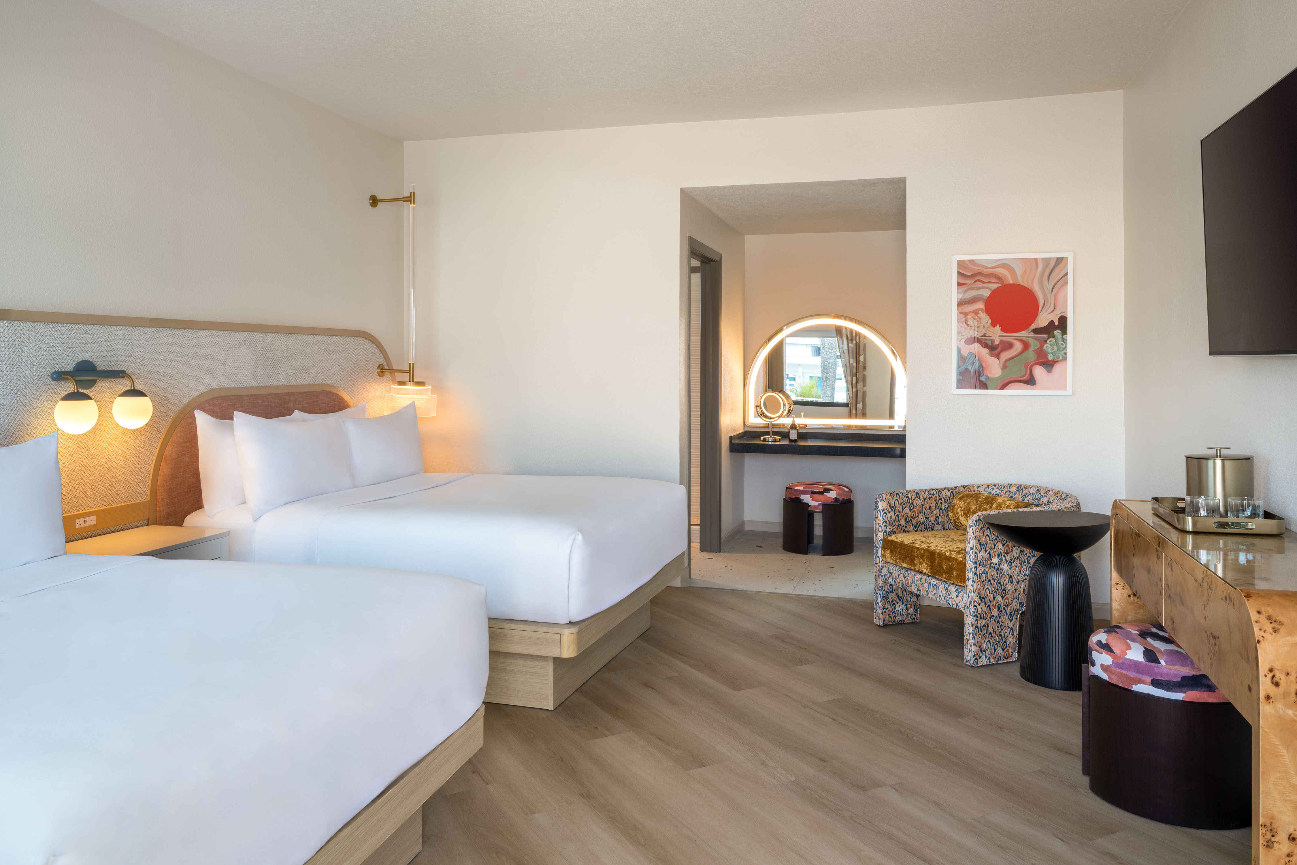 Hotel room with two queen beds, a patterned armchair, illuminated vanity mirror, and wood flooring.