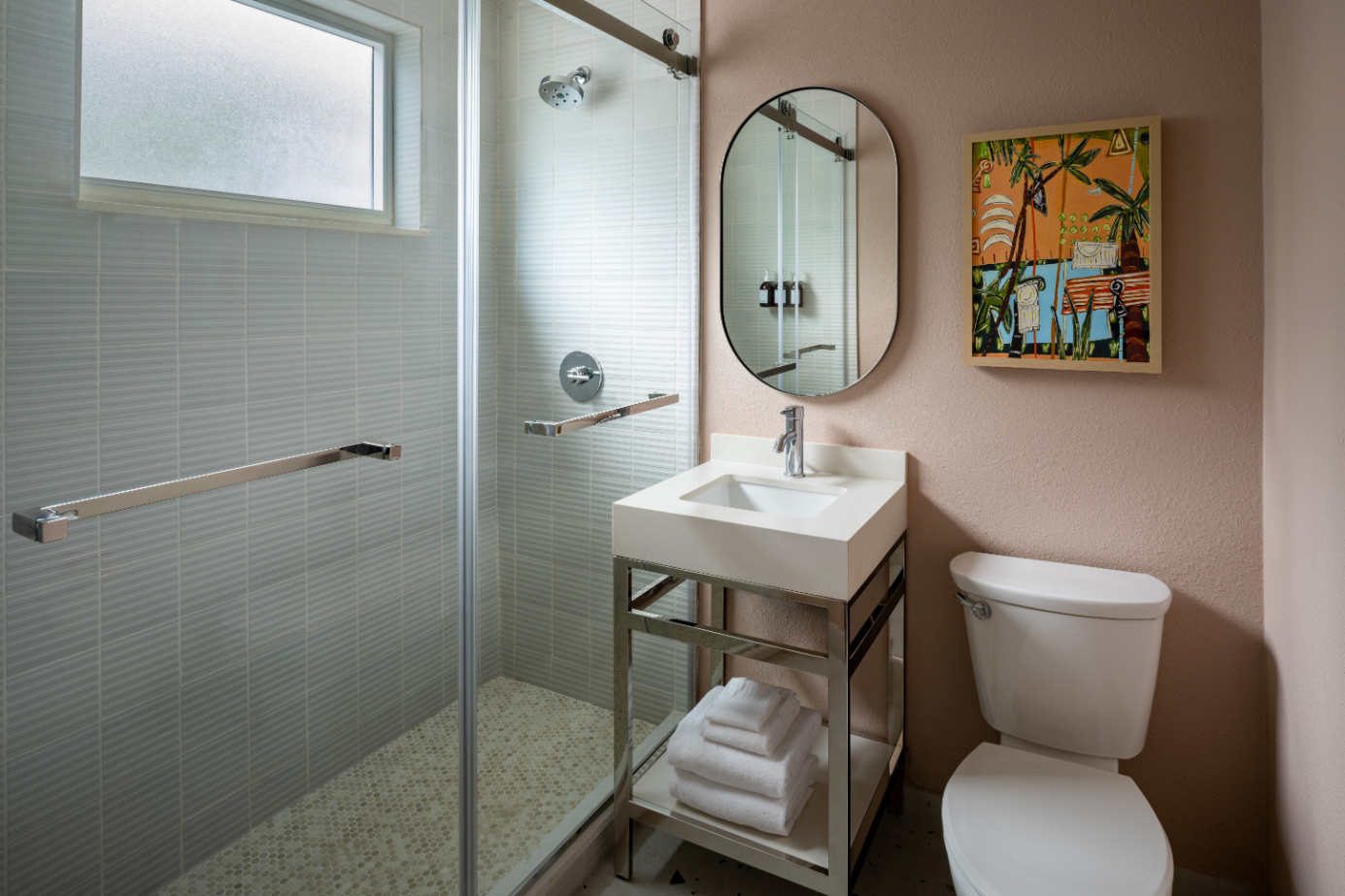 Modern bathroom with glass shower, white sink on metal stand with towels, toilet, and colorful wall art.