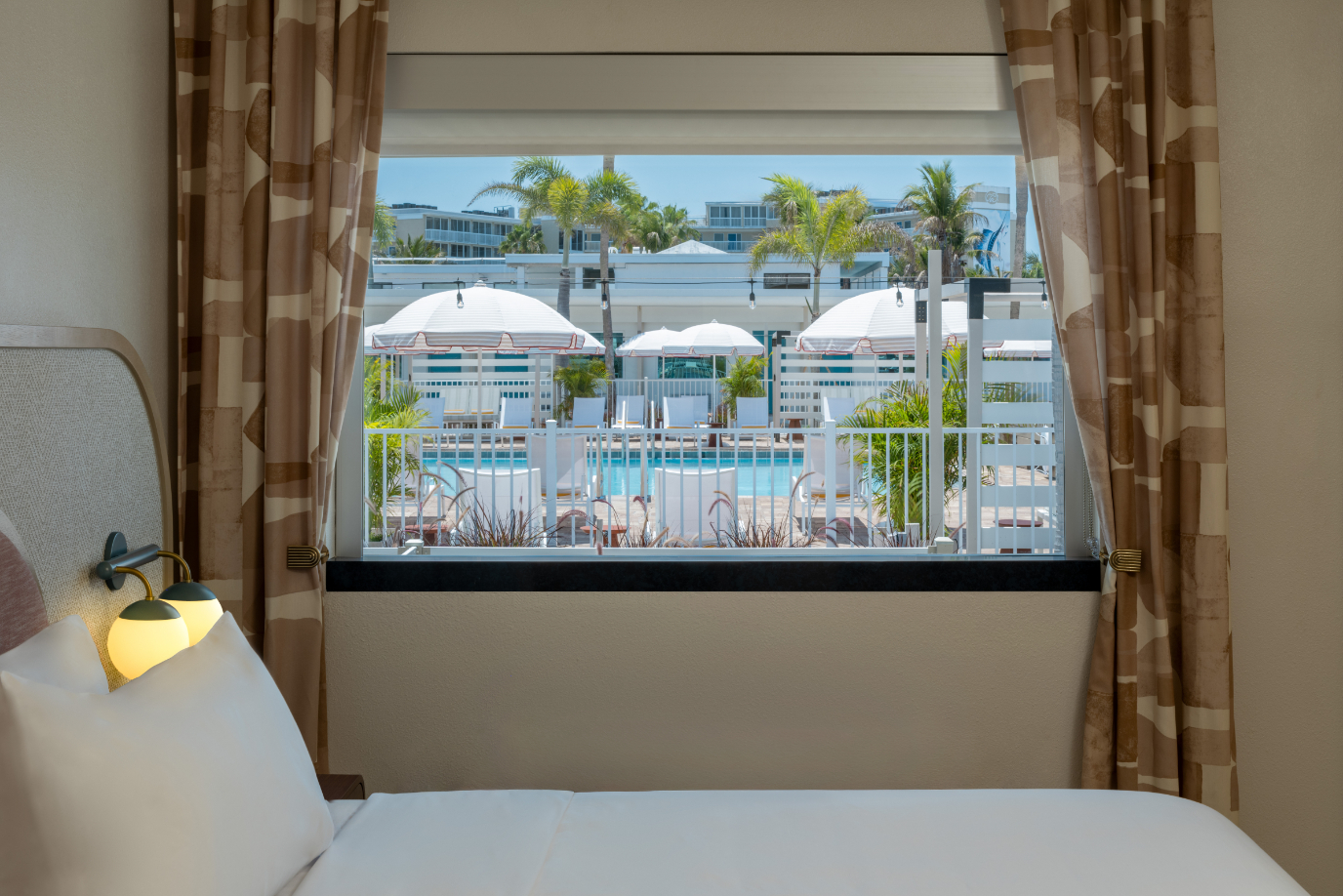Hotel room bed with view through window of pool, white umbrellas, and palm trees outside.