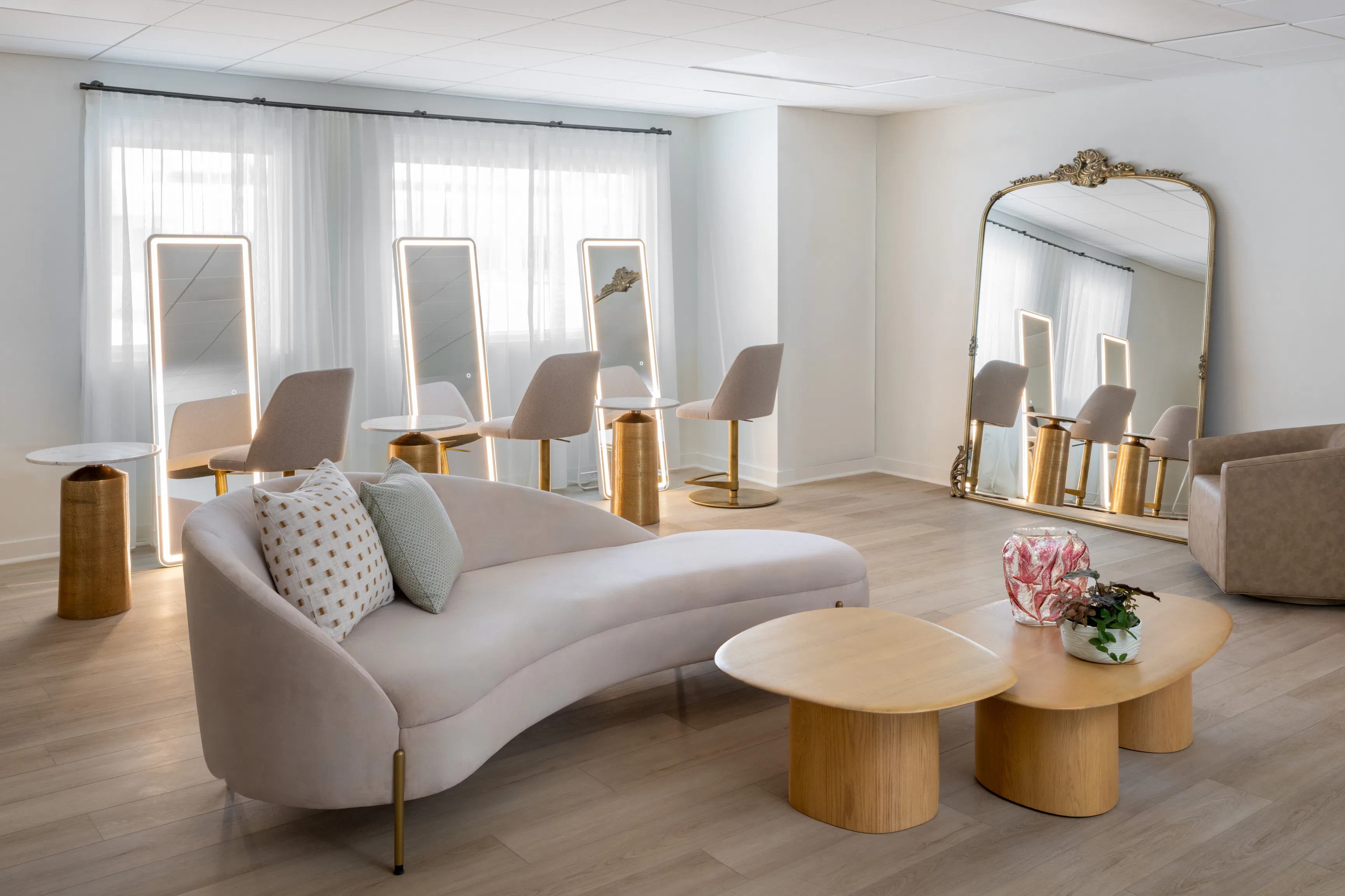 Modern room with curved pale pink sofa, wooden tables, chairs, tall lit mirrors, and large ornate wall mirror.