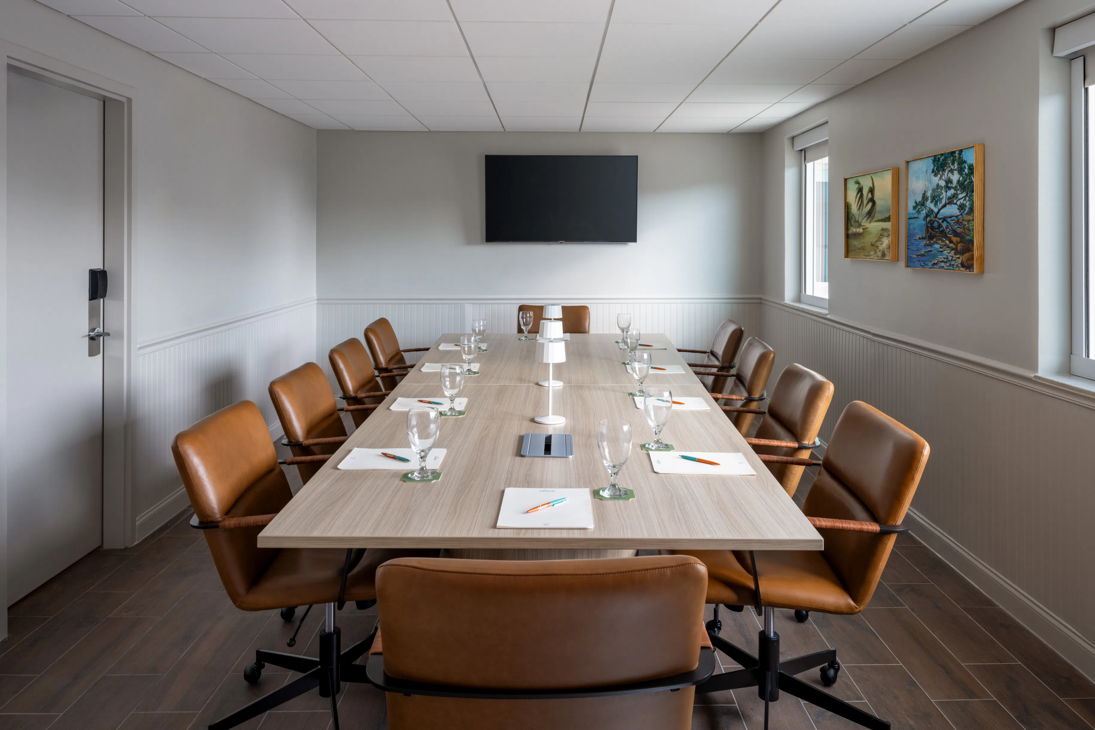 Boardroom with a long table, twelve brown leather chairs, a TV on the wall, and framed art on right wall.