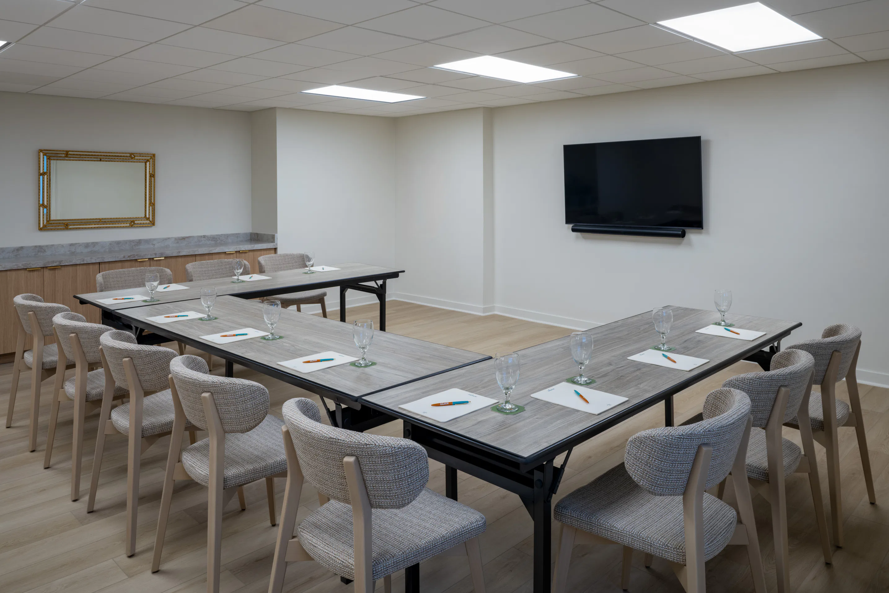 Corporate meeting room with U-shaped tables, chairs, notepads, glasses, and a wall-mounted TV screen.
