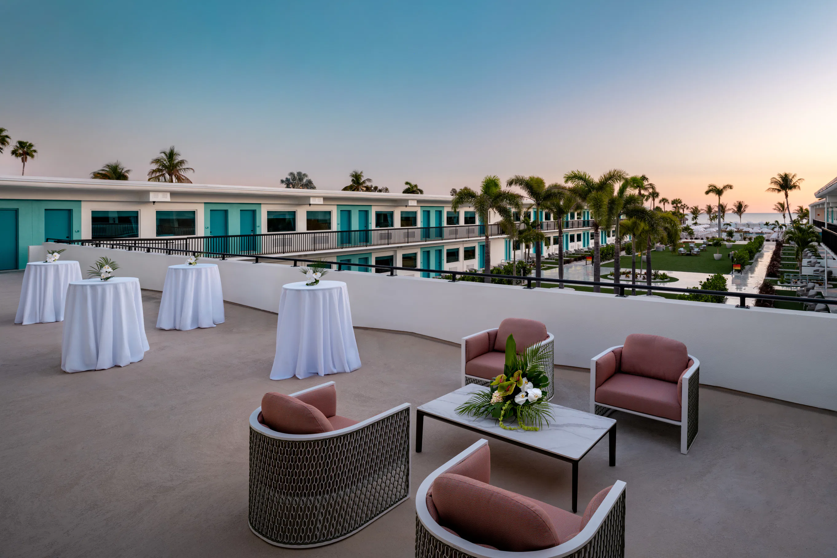 Outdoor terrace seating with pink cushioned chairs, tables with white cloths, palm trees, and ocean view at sunset.