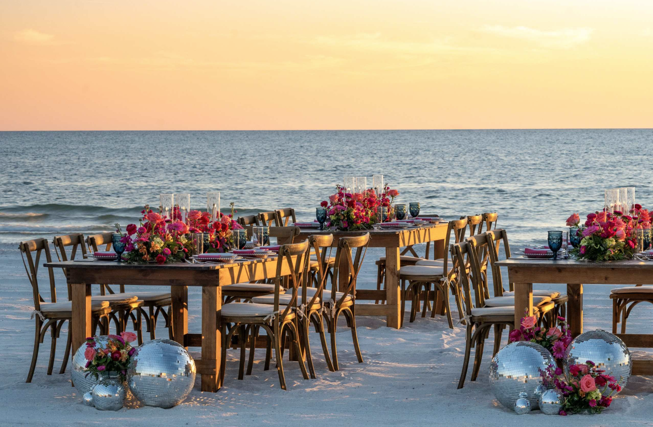 Wooden tables with floral centerpieces and disco balls on sandy beach at sunset by the ocean.