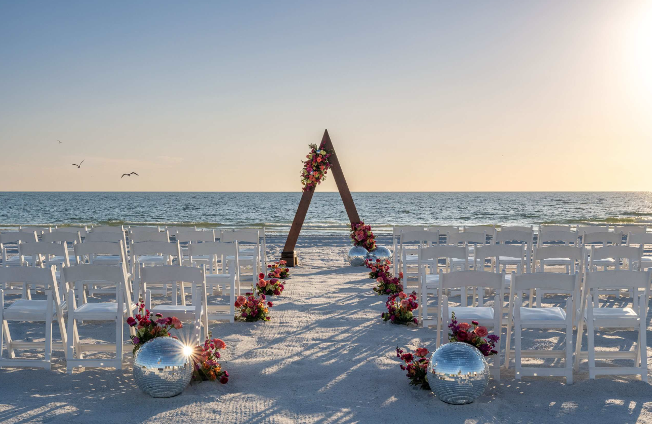 Beach wedding setup with white chairs, flower decorations, disco balls, and a wooden triangle arch by the sea.