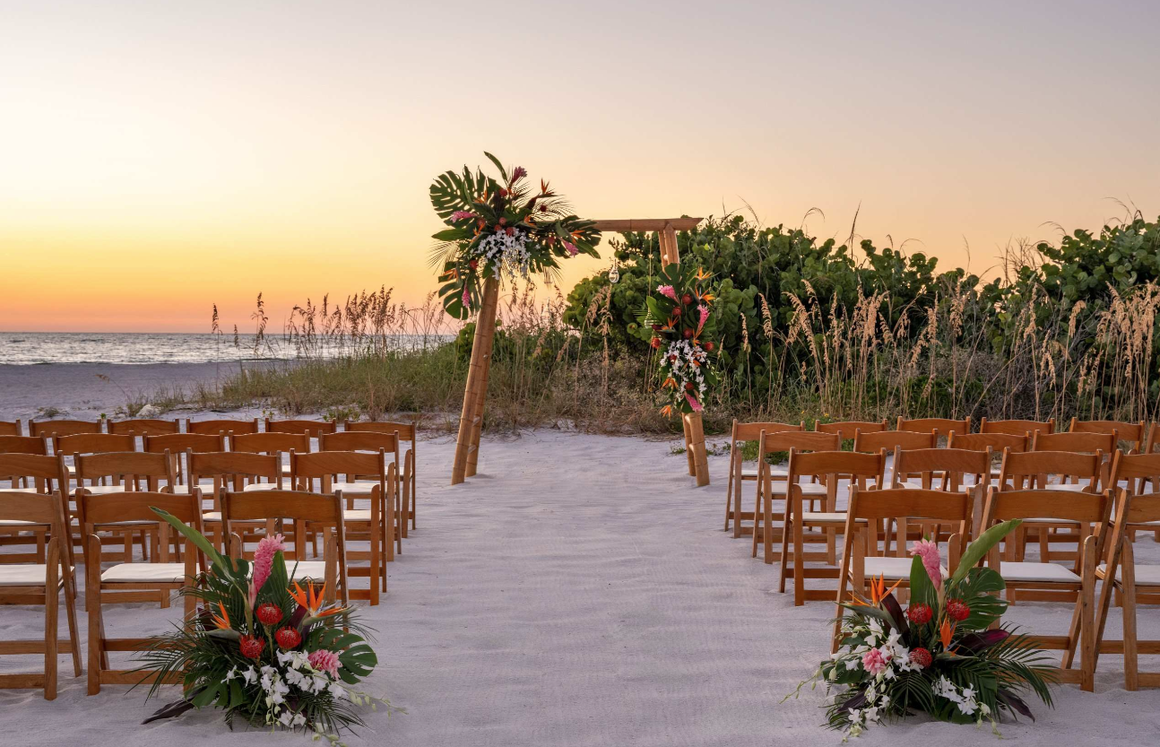 Beach wedding setup with wooden chairs, floral arrangements, and a decorated arch at sunset.