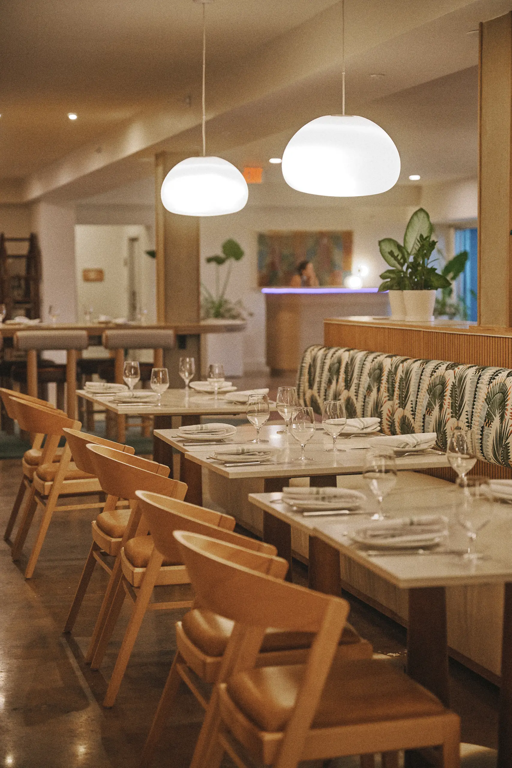 Cozy restaurant interior with wooden chairs, marble tables, patterned bench, plants, and pendant lights.