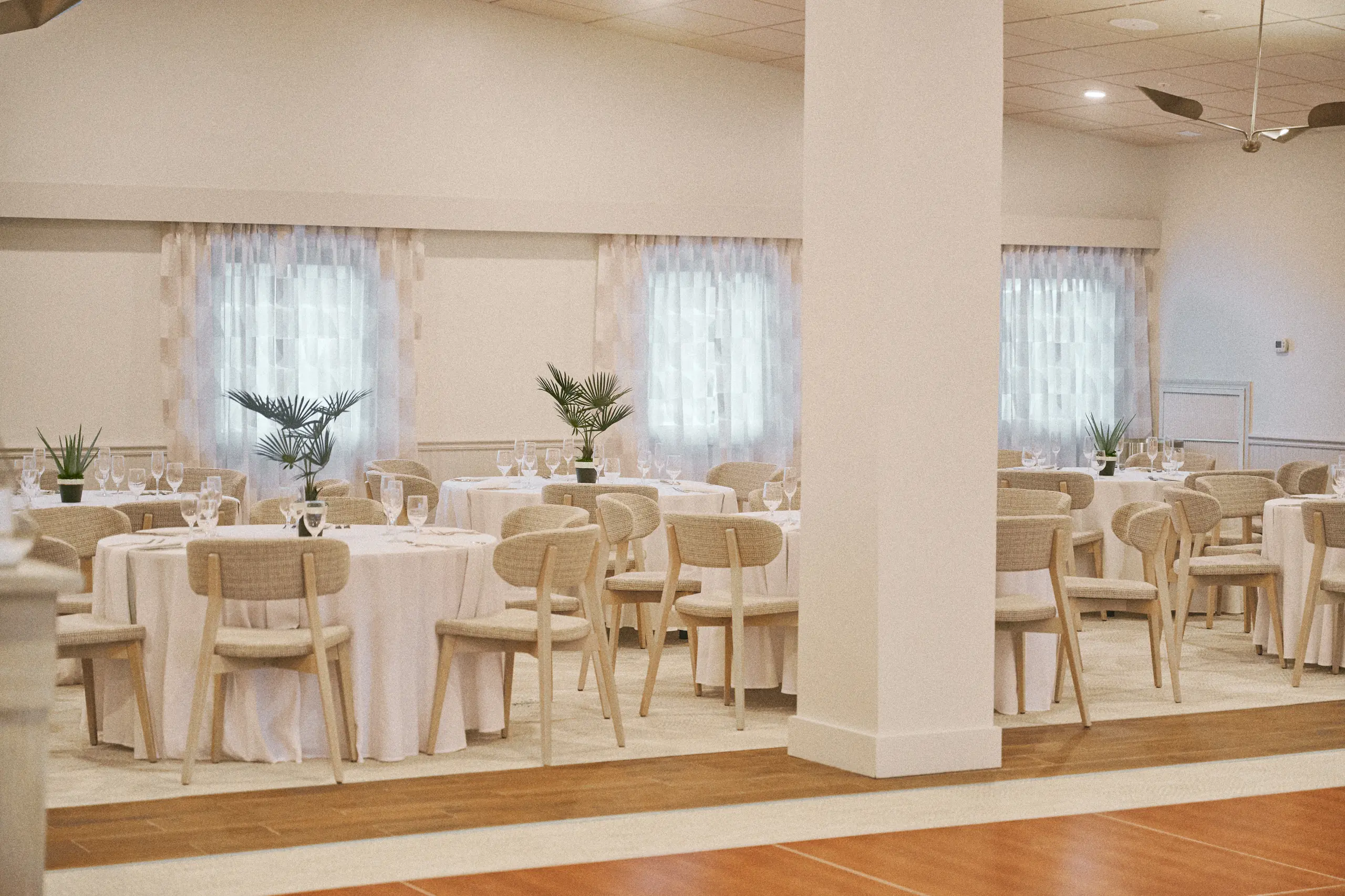 Elegant dining room with round tables covered in white linen, cushioned chairs, and potted plants.
