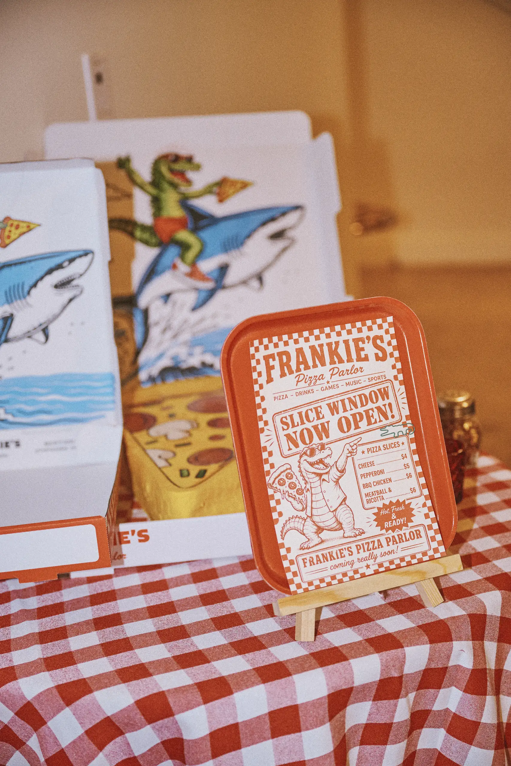 Frankie's Pizza Parlor sign on red tray with cartoon alligator holding pizza on shark in pizza boxes.