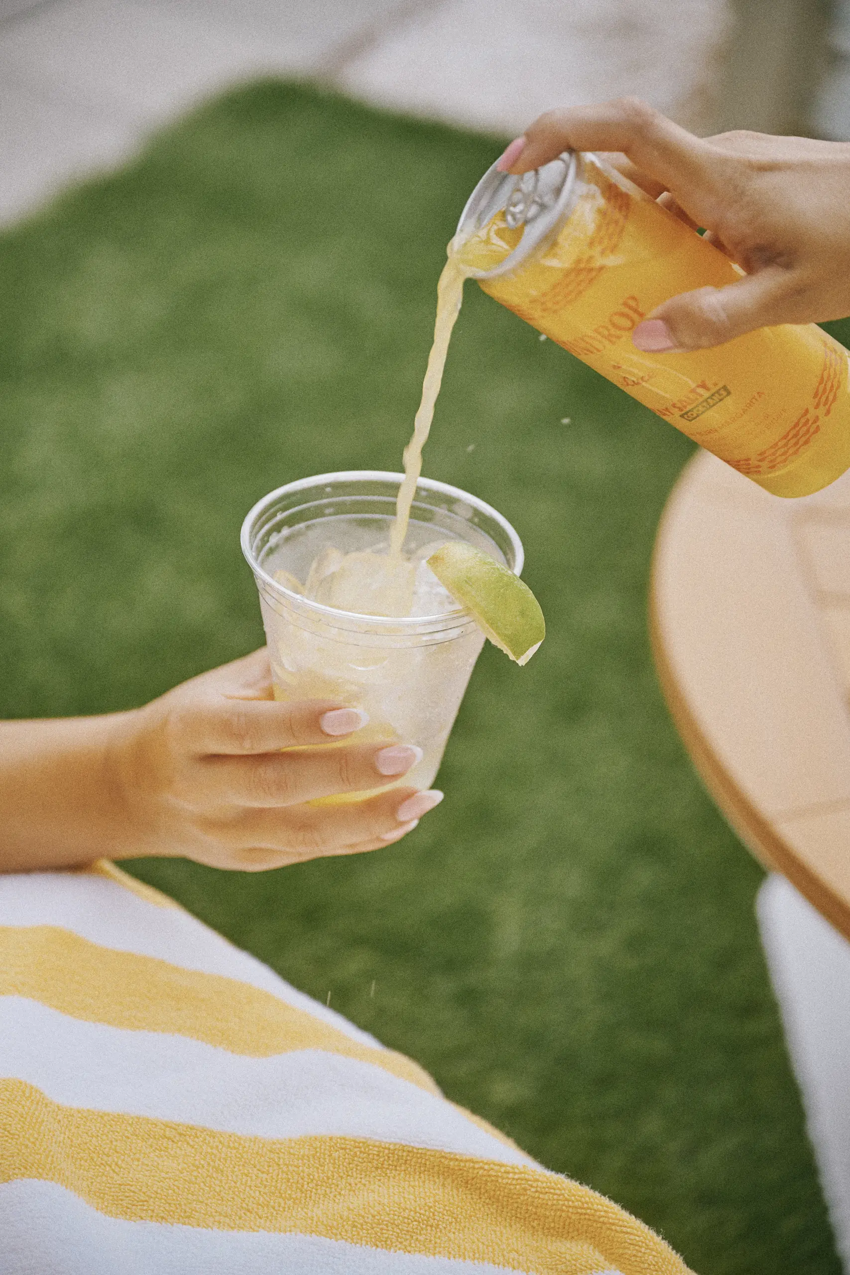 A person pours a yellow drink from a can into a cup with ice and a lime wedge outdoors.
