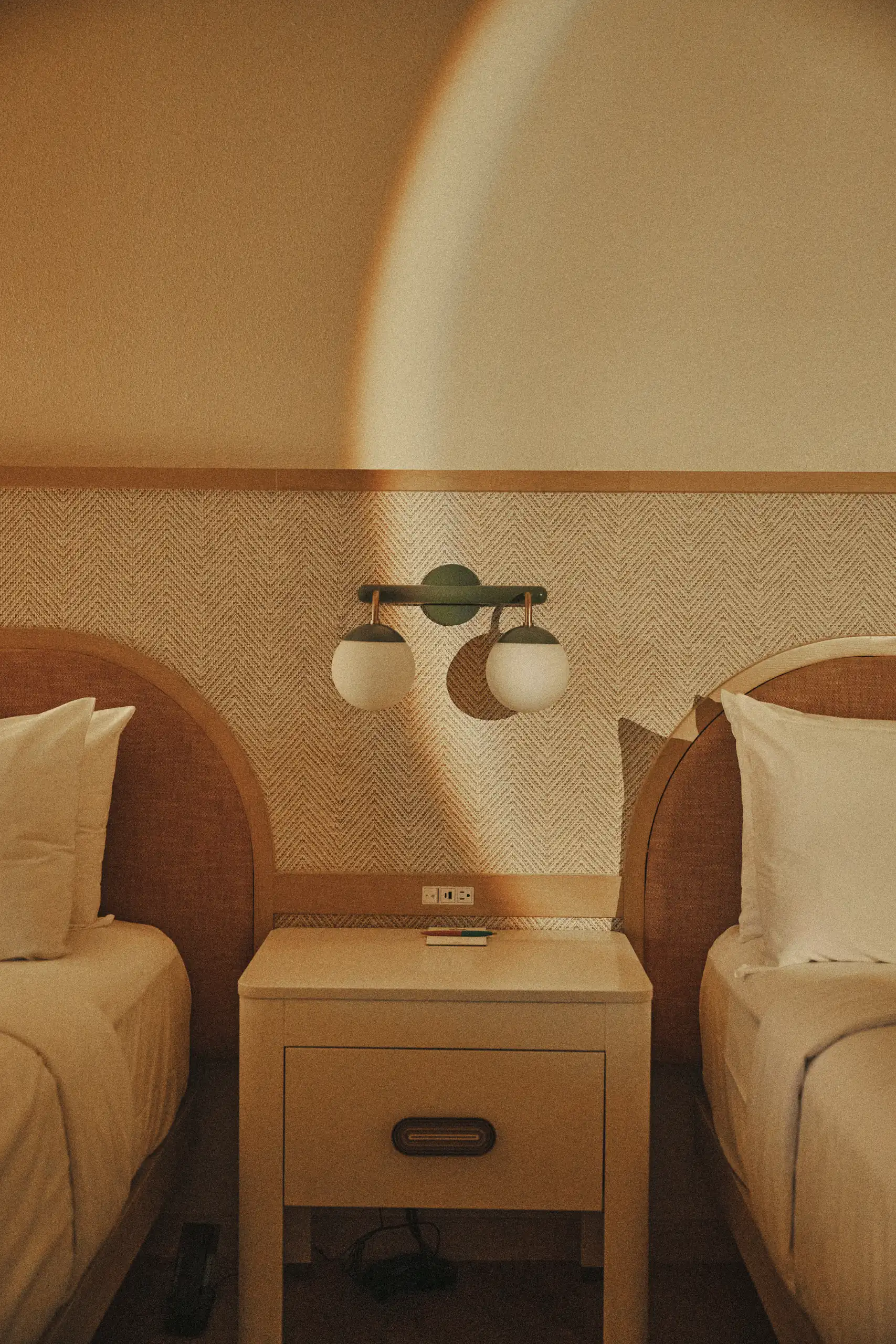 Twin beds separated by a nightstand under a wall lamp with two globe lights in warm lighting.