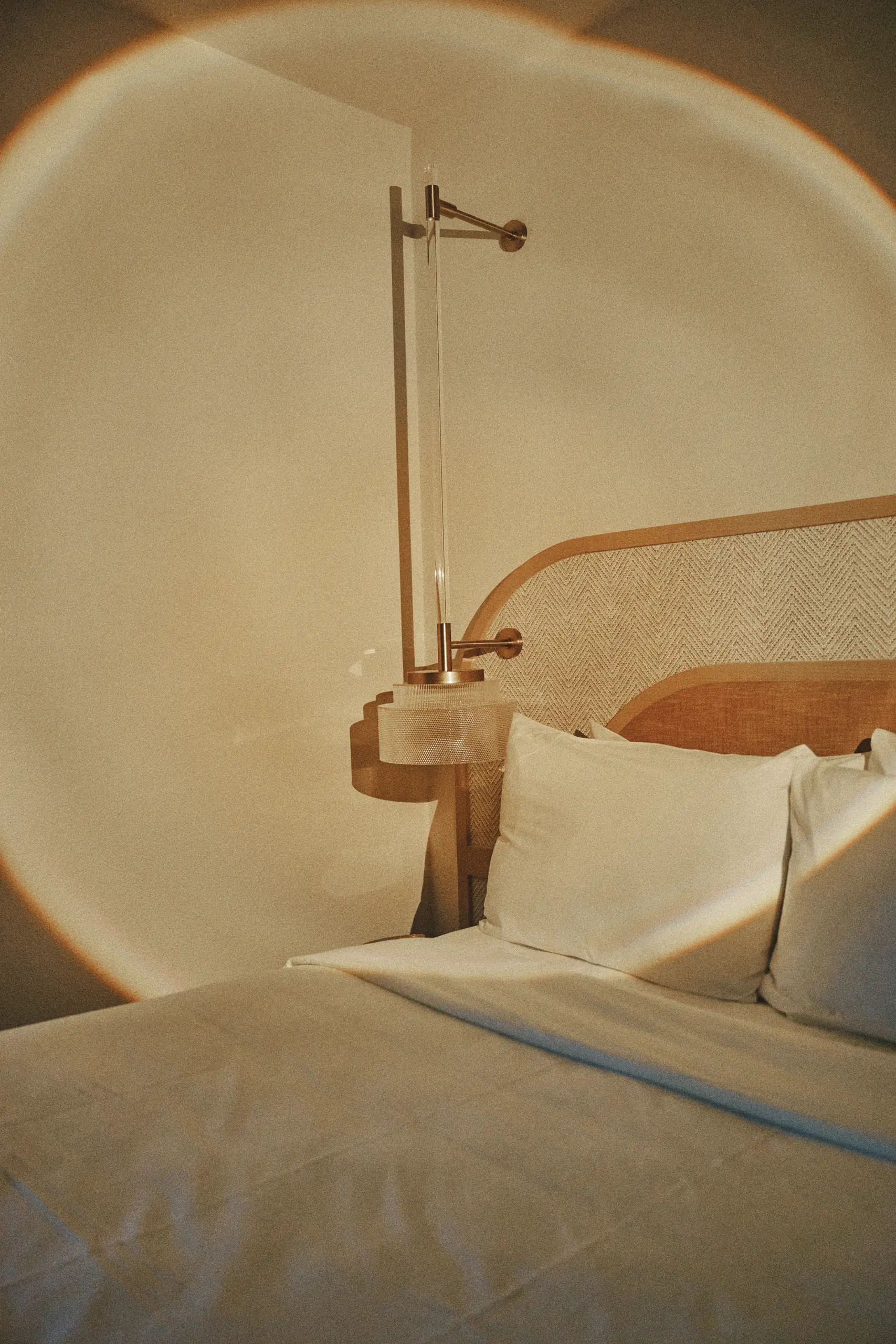 Bed with white pillows and light wood headboard under a modern wall lamp casting warm light.