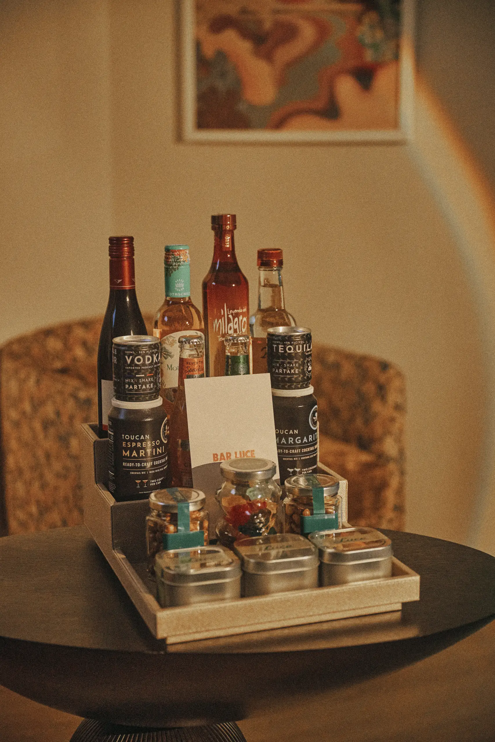 Tray on a table with assorted liquor bottles, cocktail cans, jars of nuts, and Bar Luce card.