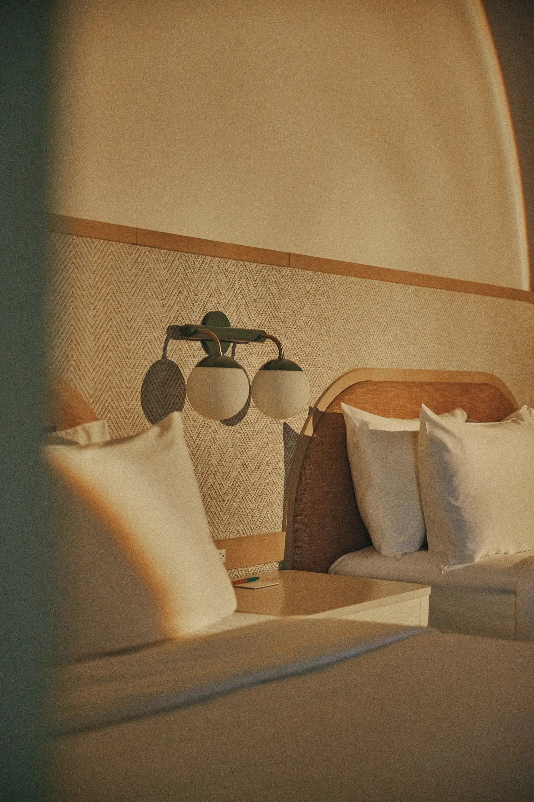 Warmly lit hotel room showing two beds with white pillows, a nightstand, and a double globe wall lamp.