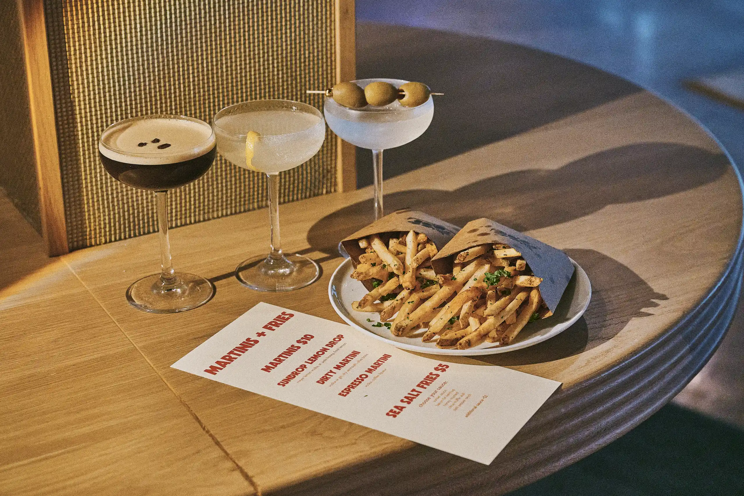 Three cocktails and two servings of seasoned fries on a wooden table with a menu titled Martinis + Fries.