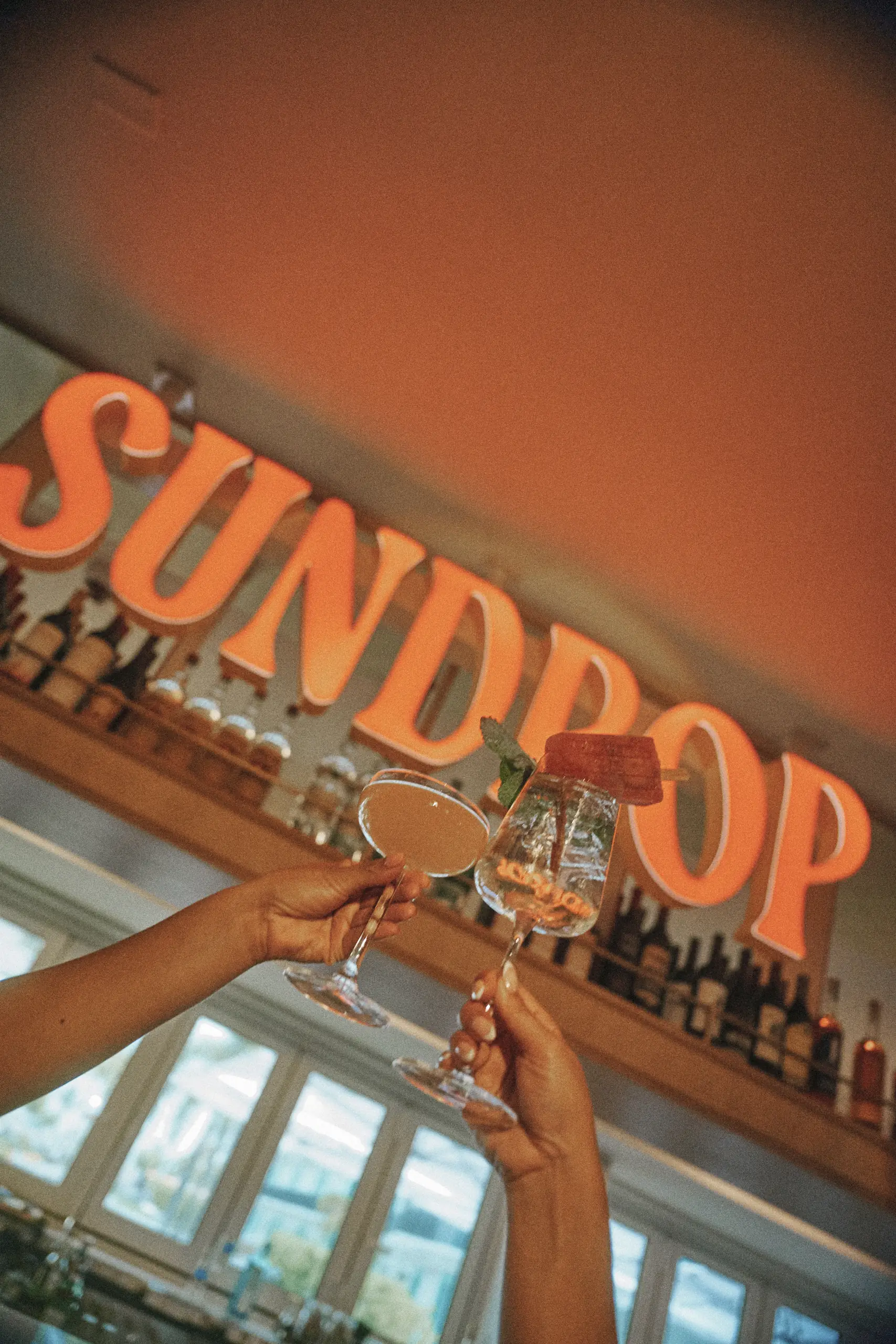 Two hands clinking cocktails in front of a glowing Sundrop sign above a bar shelf with bottles.