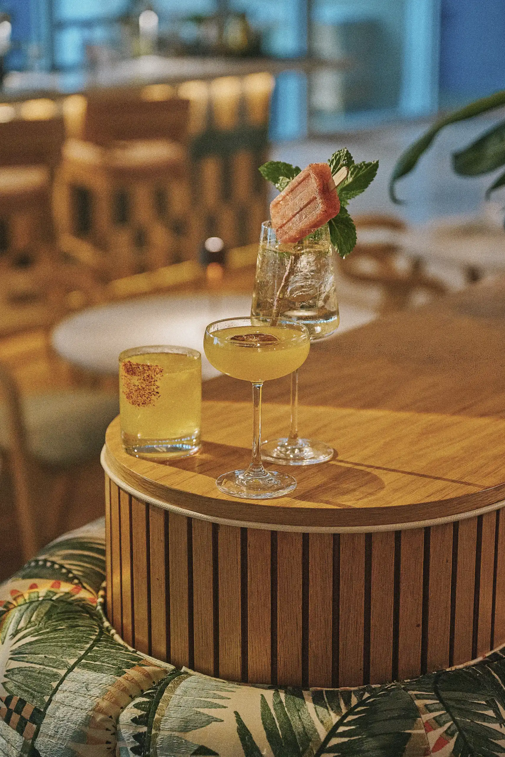 Three cocktails on a round wooden table with tropical patterned seating in a cozy bar setting.