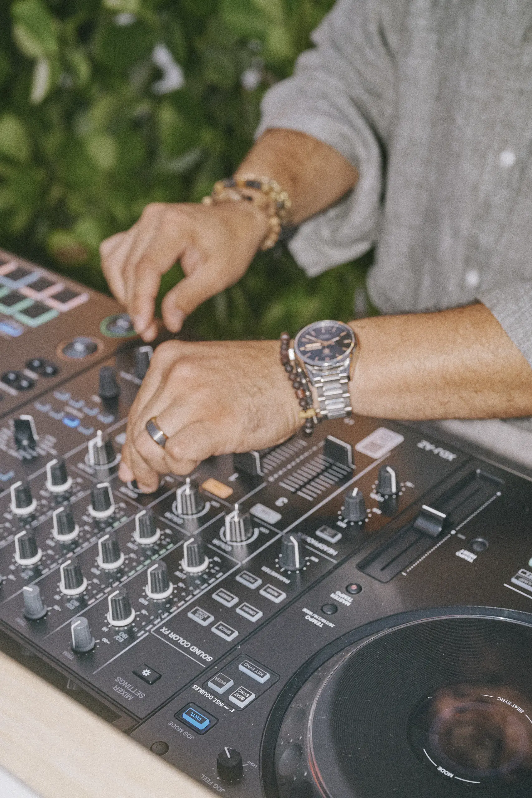 Hands adjusting knobs on a DJ mixer with buttons and sliders, leafy green background.