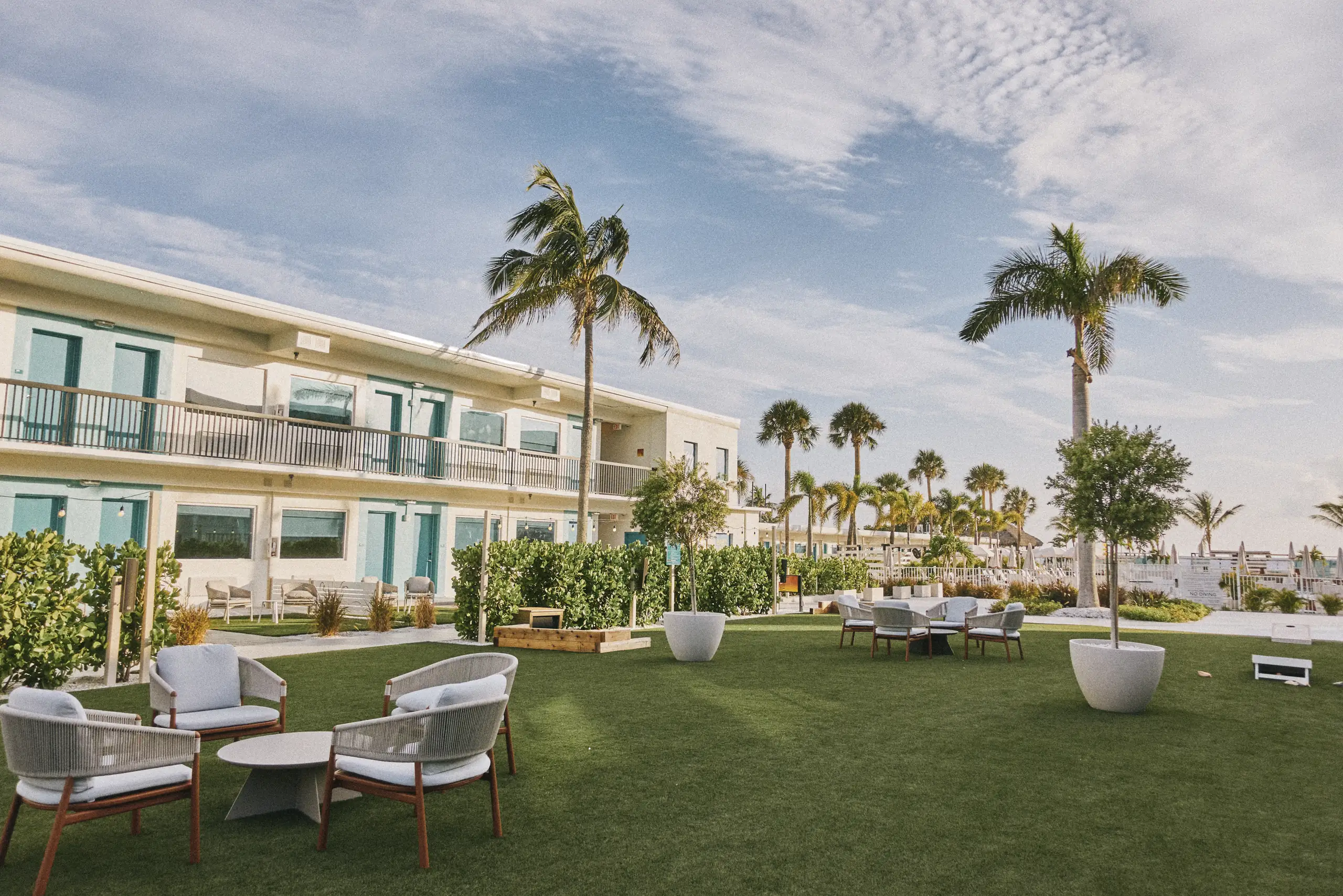 Outdoor seating area with chairs and tables on green lawn near a building and palm trees under blue sky.