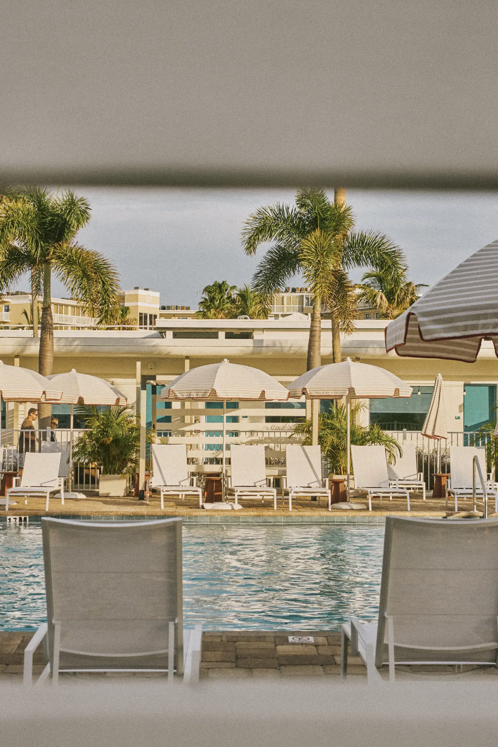 View of a swimming pool with white lounge chairs and umbrellas, framed by window blinds.