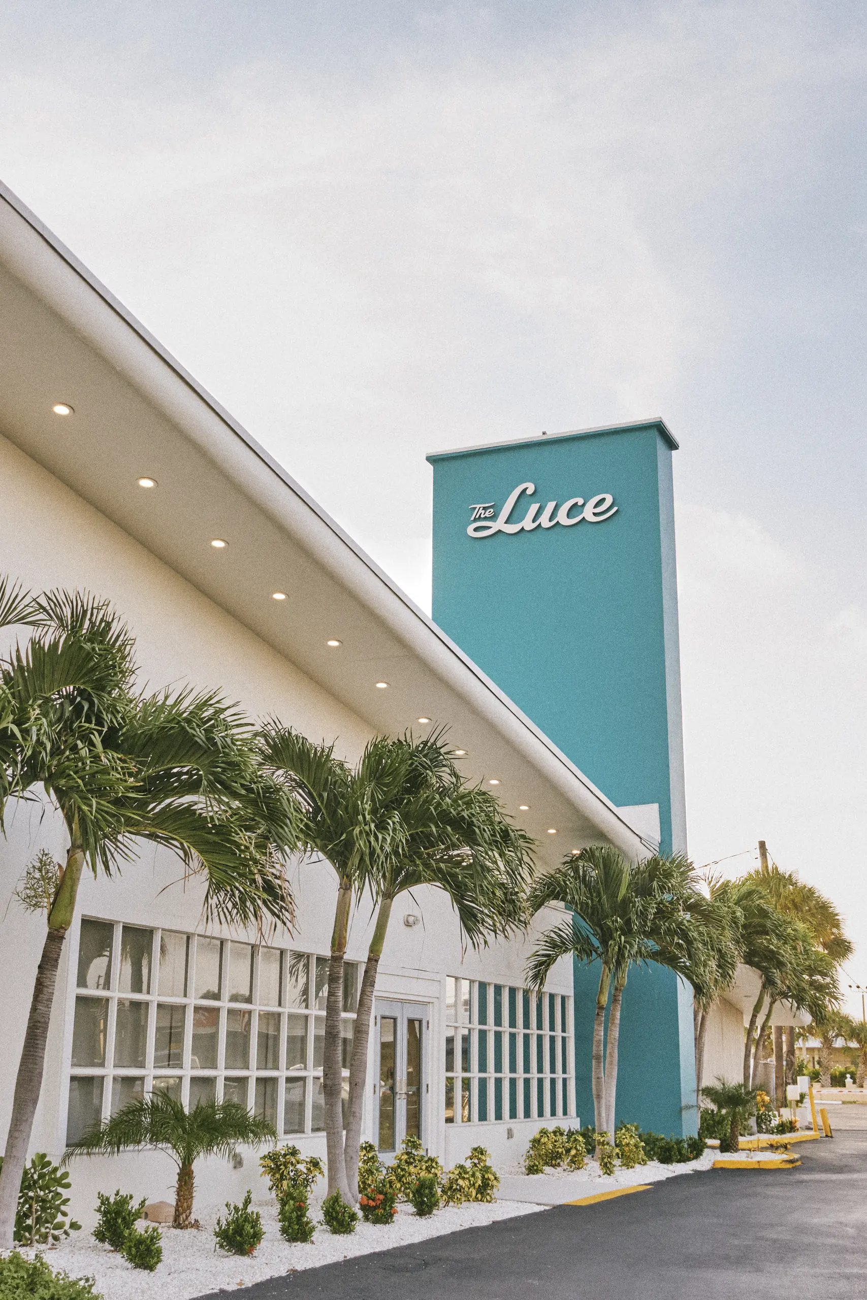 White building with teal tower labeled The Luce, lined with palm trees and plants in front.