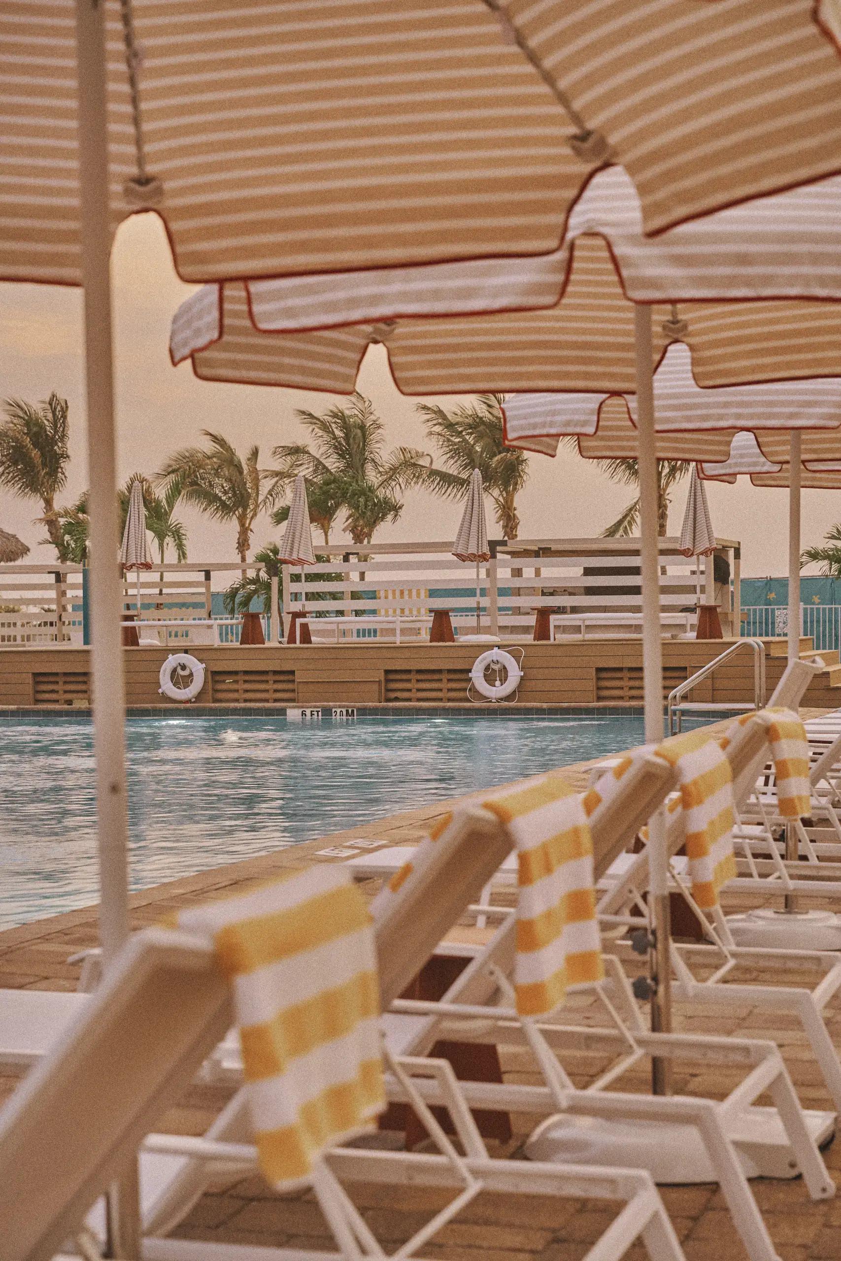 Poolside lounge chairs with yellow-striped towels under striped umbrellas near a pool and palm trees.