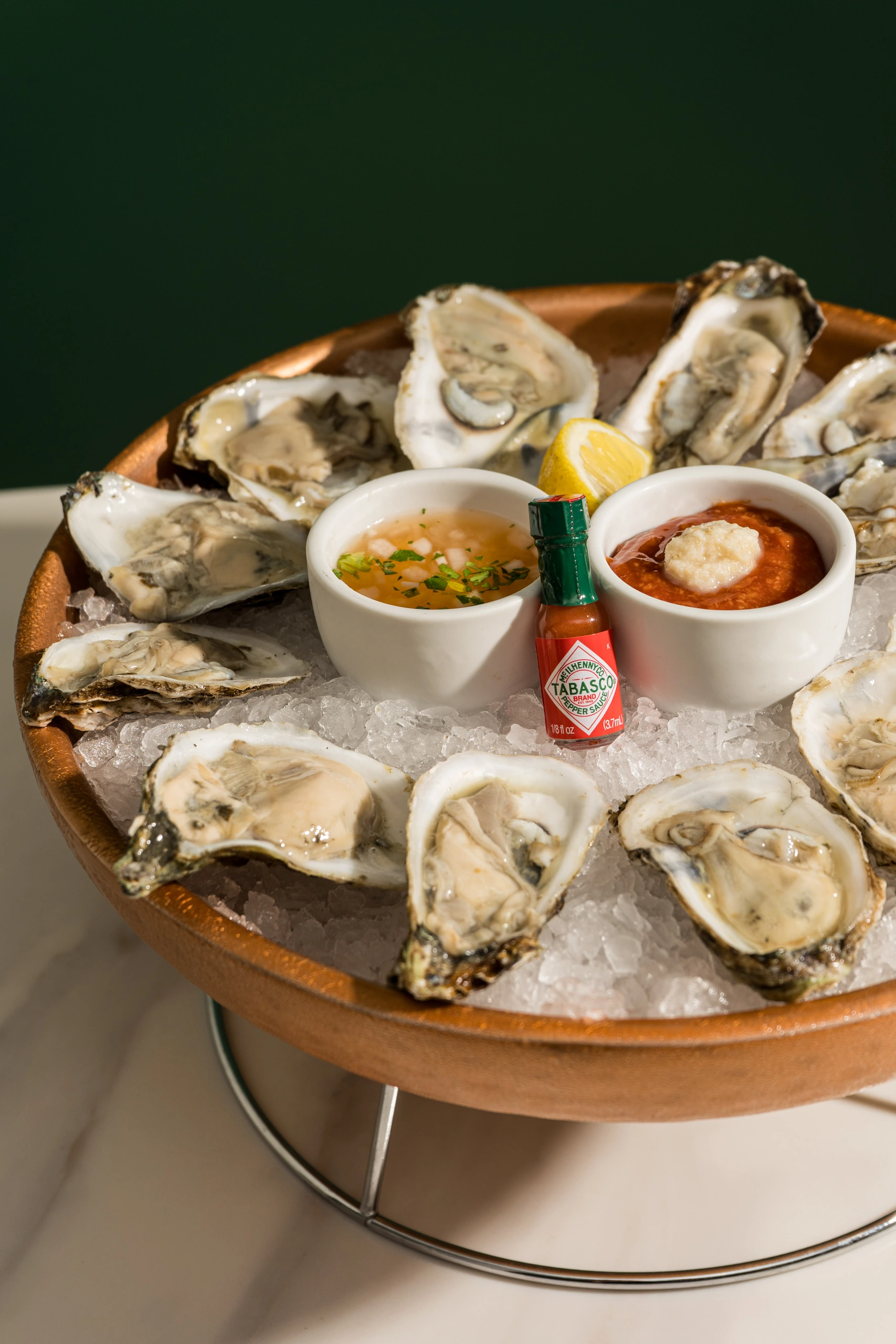 Oysters on ice with lemon, Tabasco sauce, and two dipping sauces in a brown bowl on a metal stand.