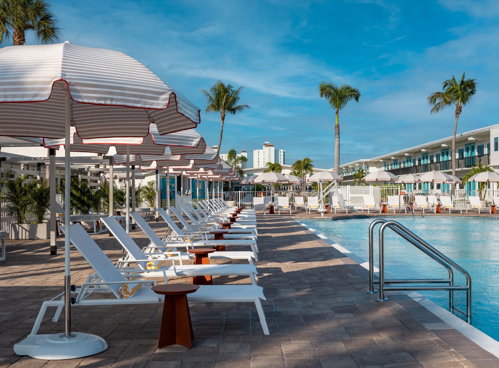 Empty poolside with white lounge chairs, striped umbrellas, palm trees, and clear blue sky at a resort.