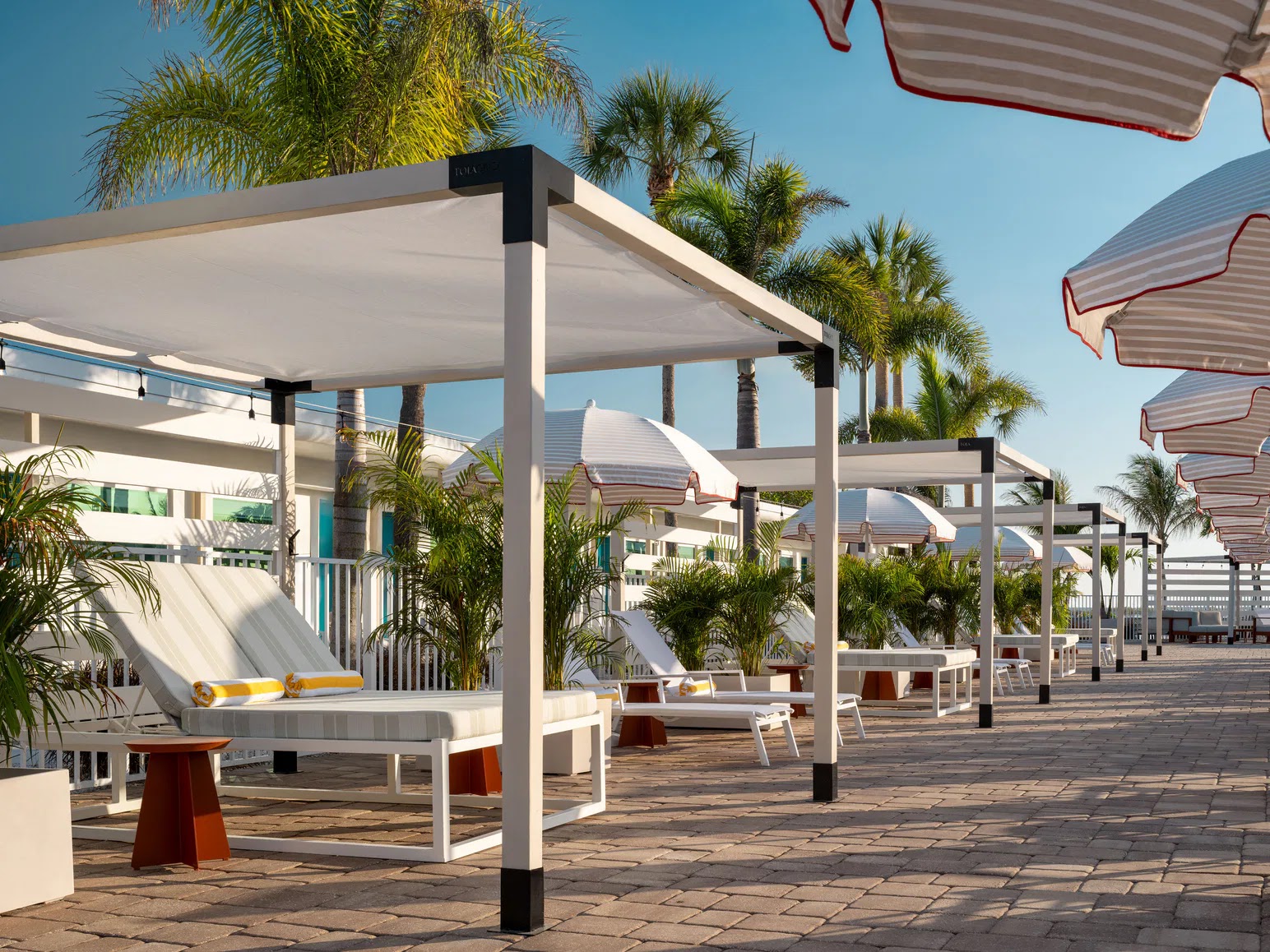 Row of white poolside cabanas with lounge chairs, umbrellas, and palm trees under clear blue sky.