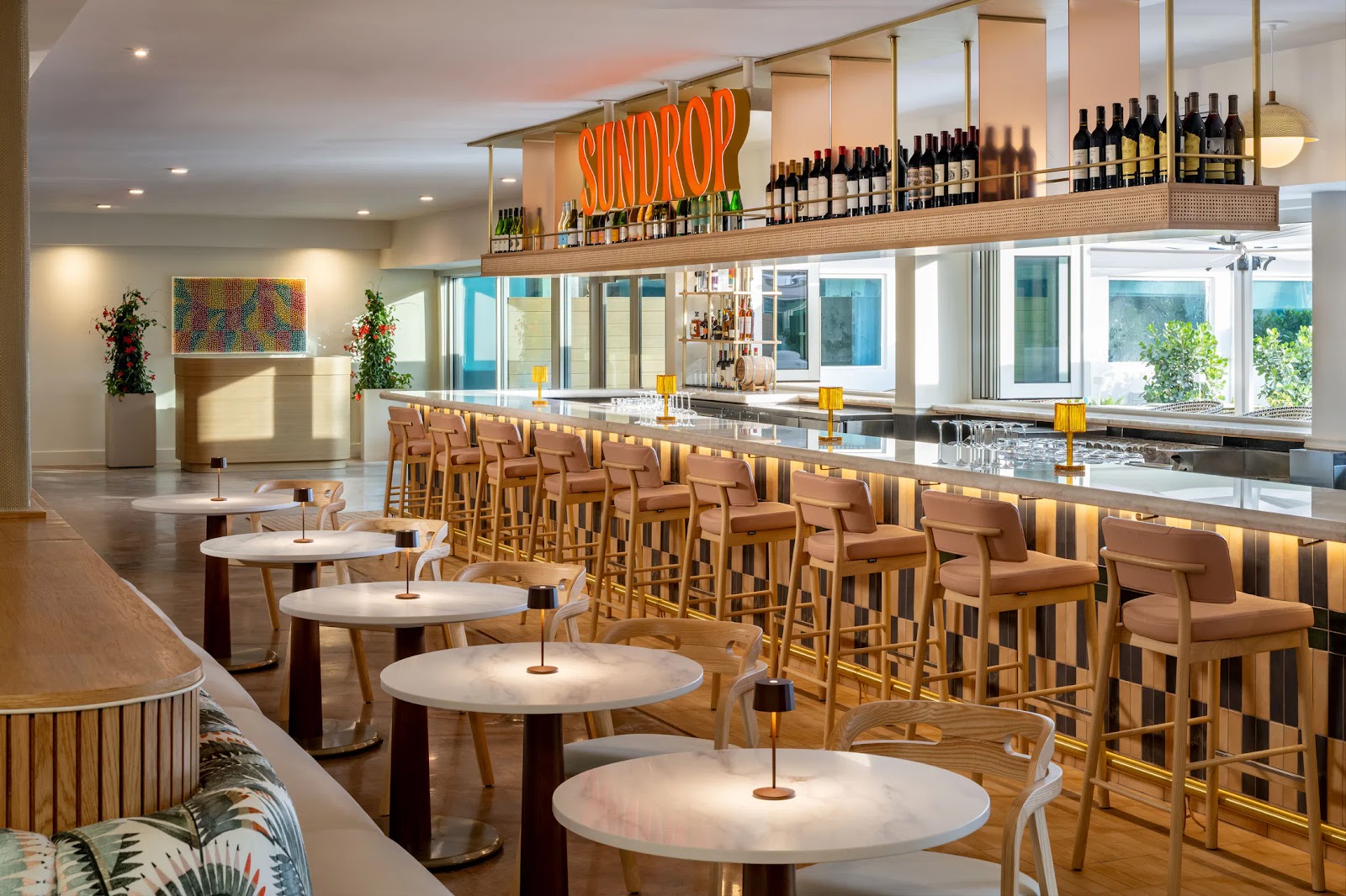 Bright bar area with cushioned stools, round tables, small lamps, and 'SUNDROP' sign above wine bottles.