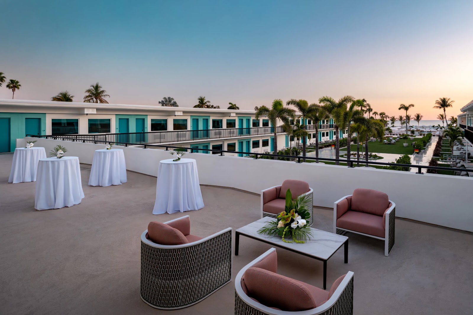 Outdoor terrace with seating and tables overlooking a hotel courtyard with palm trees at sunset.