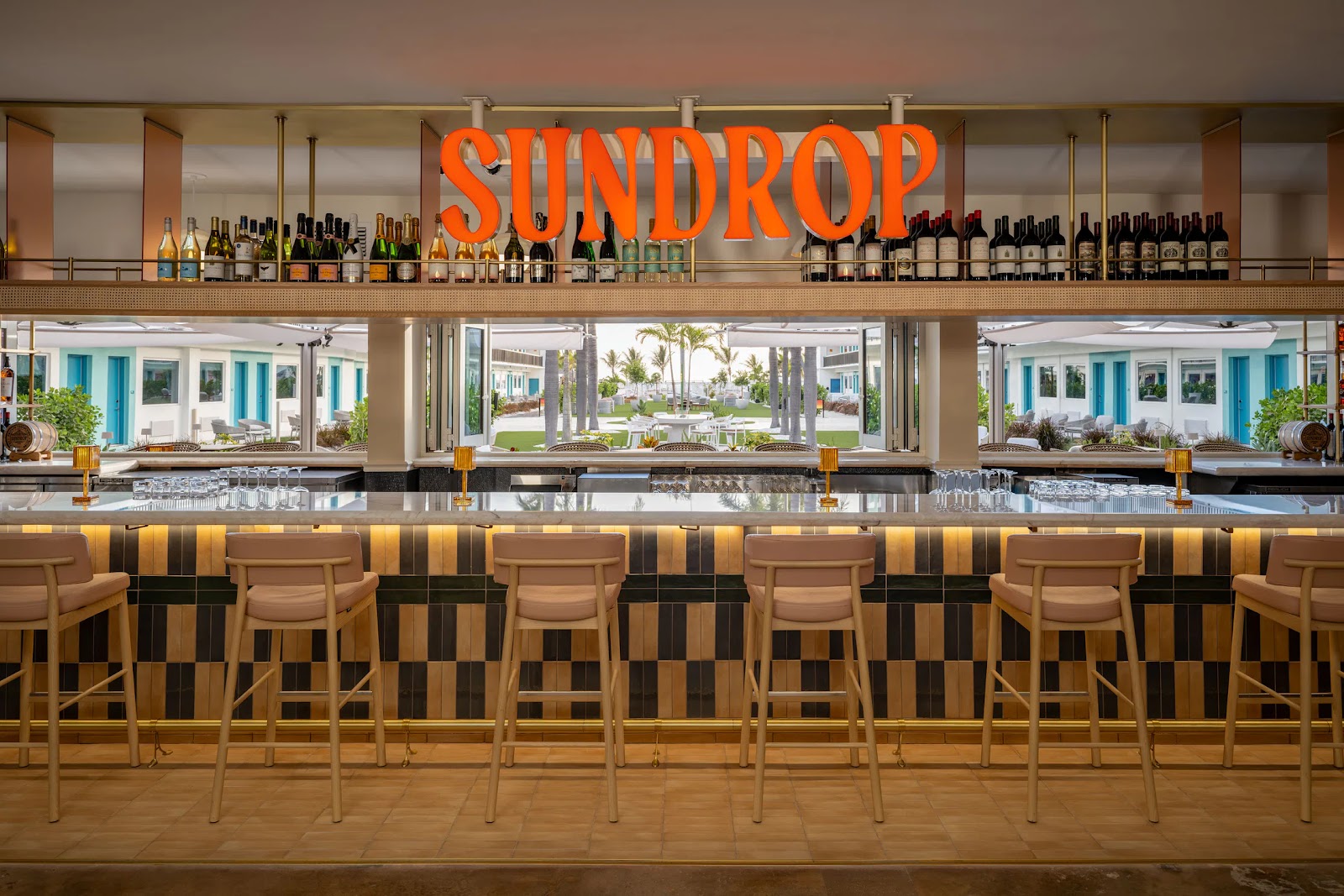 Modern bar with tan stools and bottles, large SUNDROP sign, looking out to palm trees and outdoor seating.